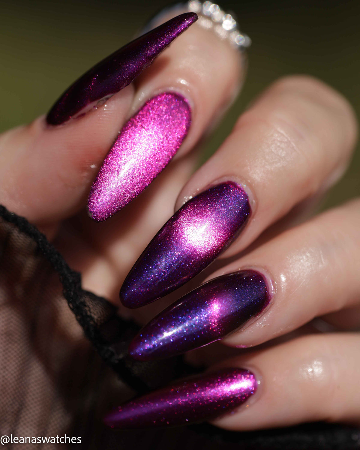 A close-up of a hand with long stiletto-shaped nails painted in a metallic magenta-to-purple iridescent polish, with a ring visible on one finger against a blurred olive background.