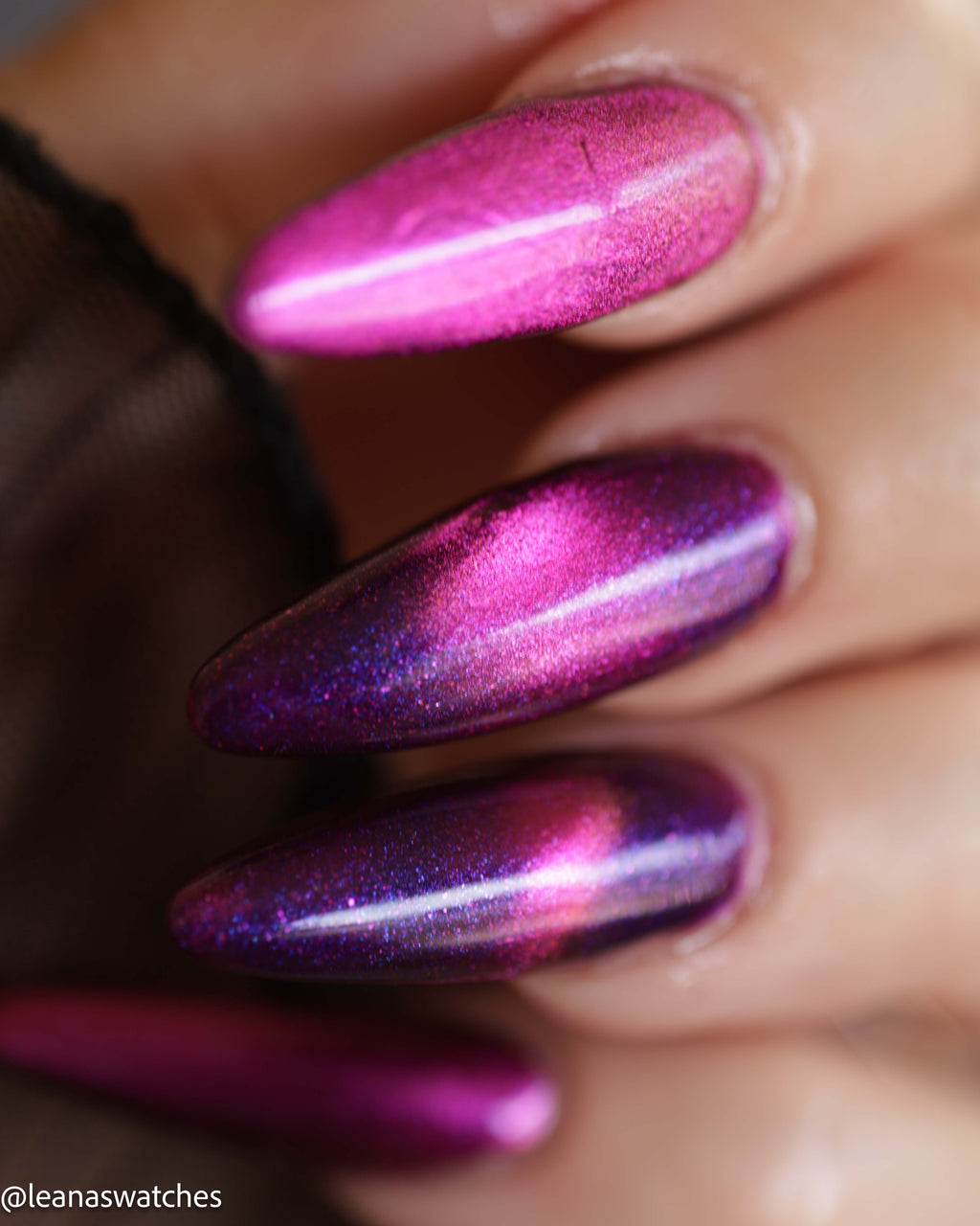 A close-up of a hand with long, almond-shaped nails painted in a metallic magenta and purple glitter nail polish that shifts under light.