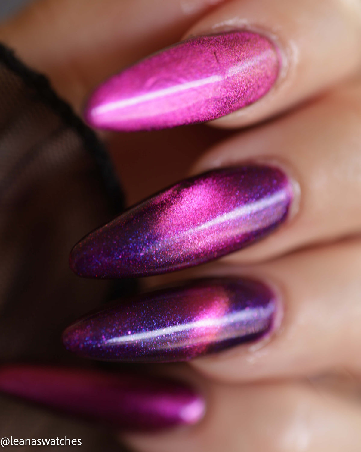 A close-up of a hand with long, almond-shaped nails painted in a metallic magenta and purple glitter nail polish that shifts under light.