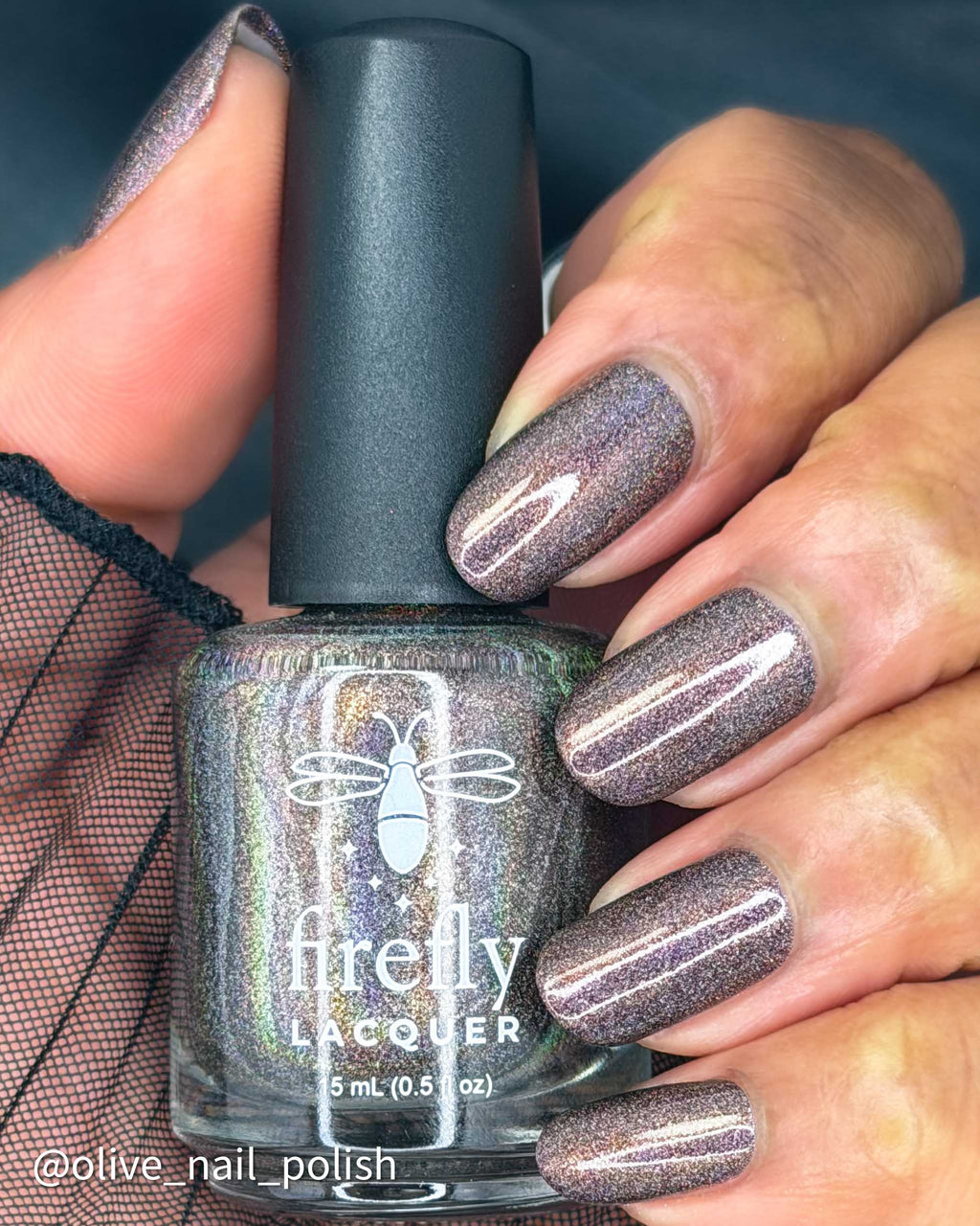 A close-up of a hand holding a bottle of Firefly Lacquer nail polish. The nails are painted with a shimmering, metallic lavender-gray polish, and the bottle itself is a dark smoky gray with a holographic finish showing green and gold iridescence.