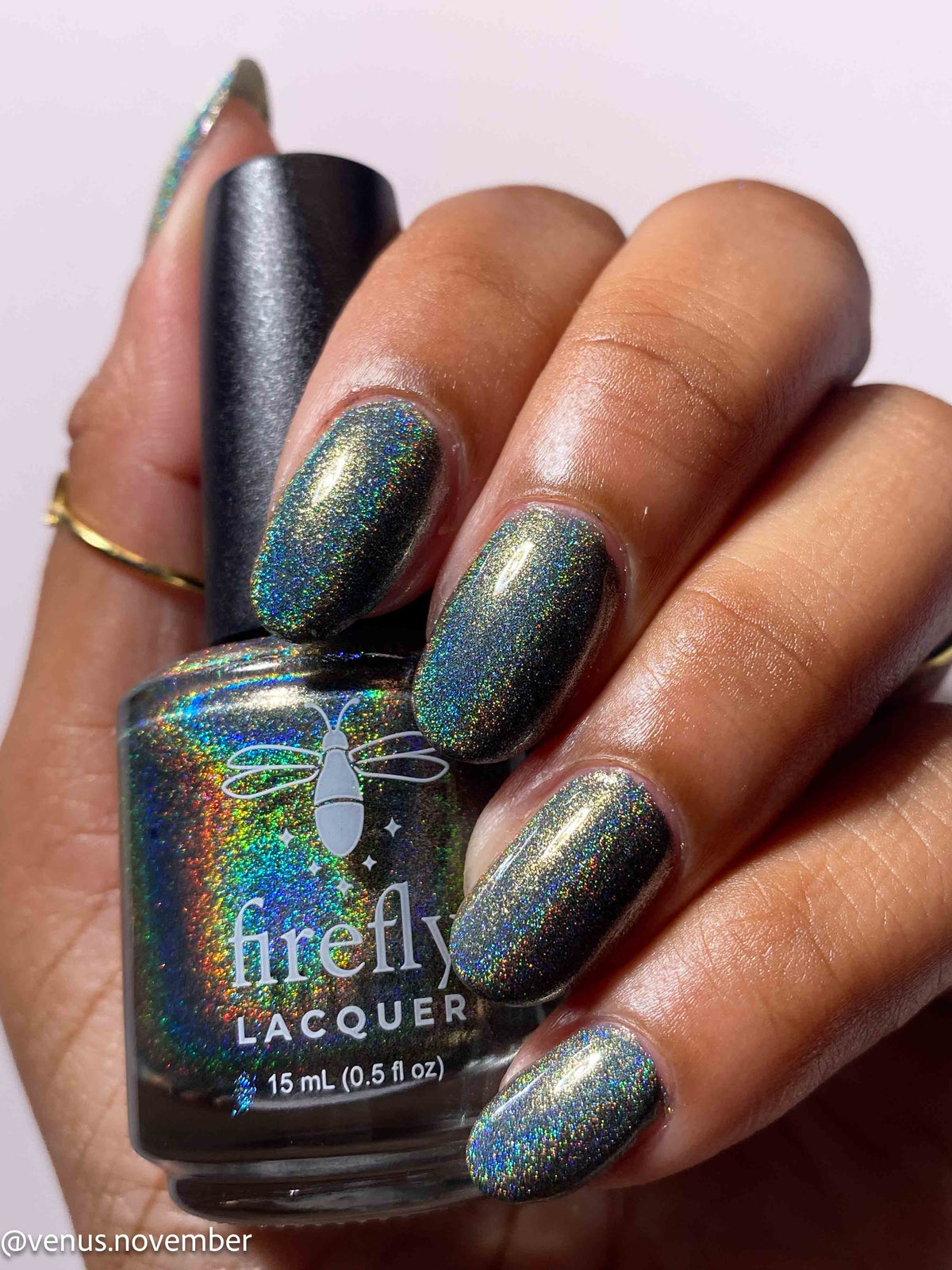 A hand with nails painted in a shimmering holographic nail polish, holding a bottle of Firefly Lacquer in the same shade. The polish has an iridescent, holographic finish reflecting green, blue, and gold tones.