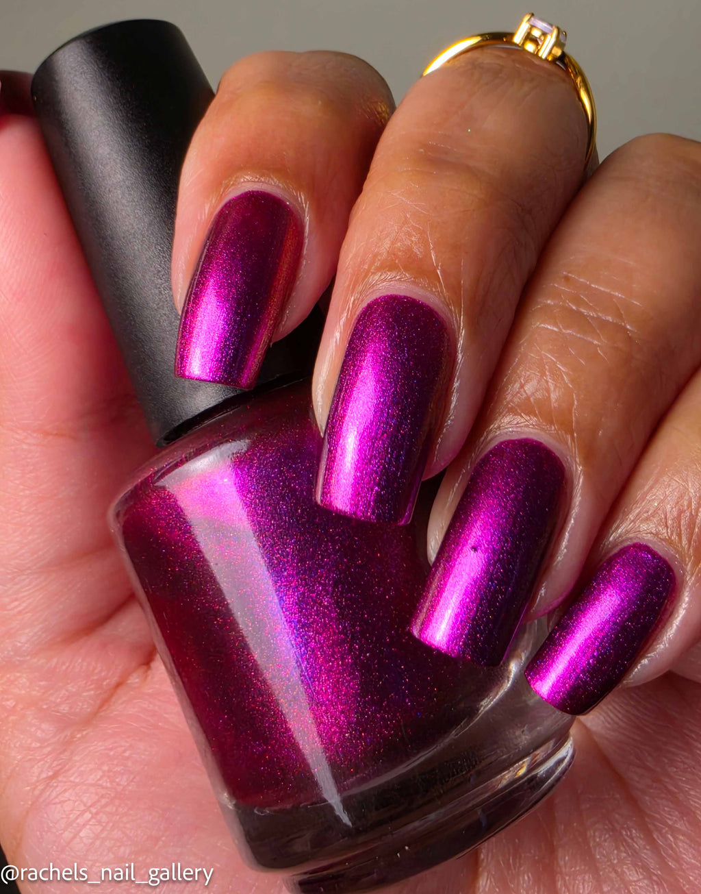 A close-up of a hand holding a bottle of metallic magenta nail polish, with the nails painted in the same shimmering purple-pink shade. The bottle has a black cap, and a gold ring is visible on one finger.