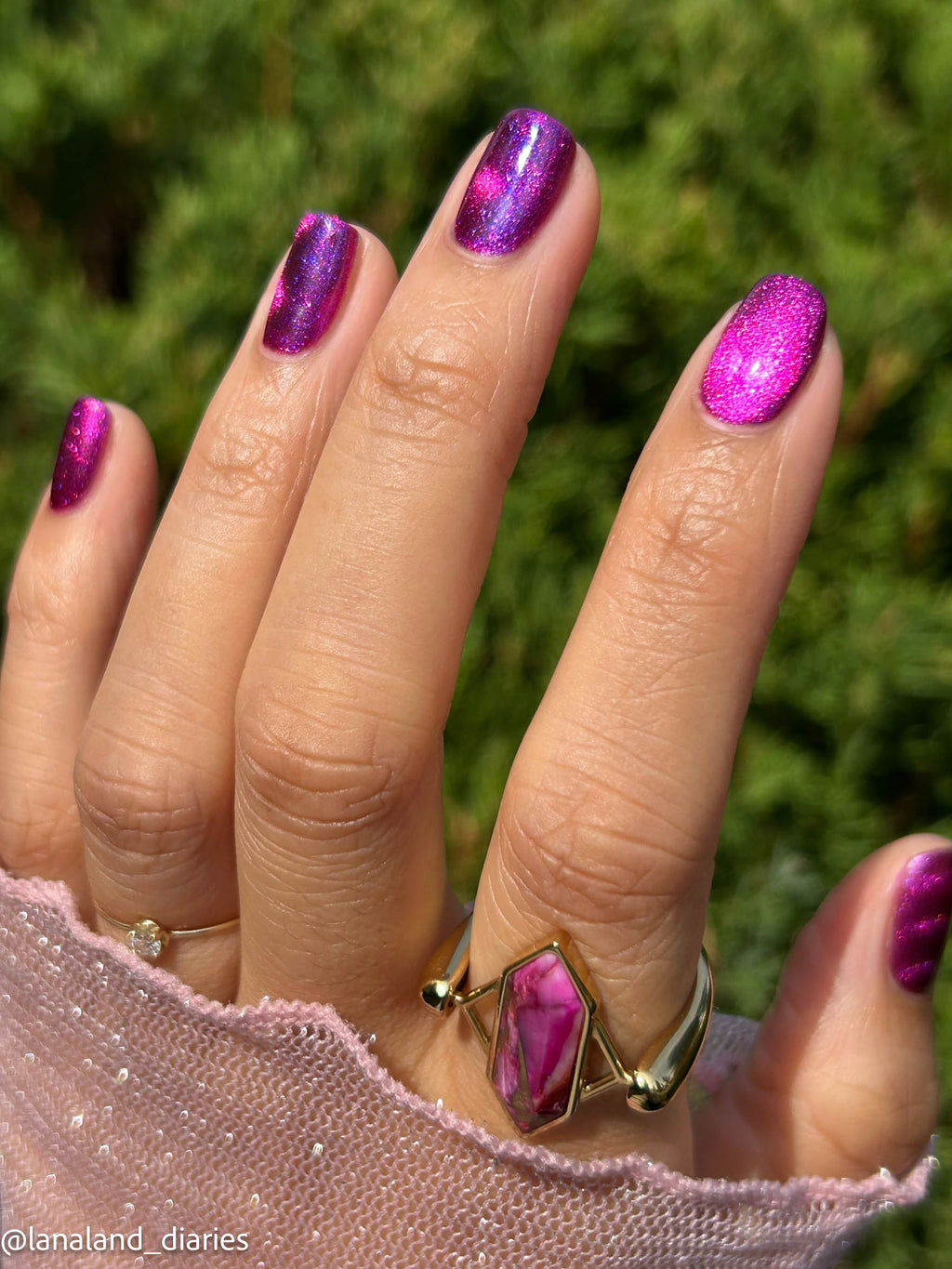 A close-up of a hand with fingernails painted in a sparkling magenta-purple cat-eye polish, wearing a gold ring with a pink stone. The background is a soft-focus green outdoor setting.