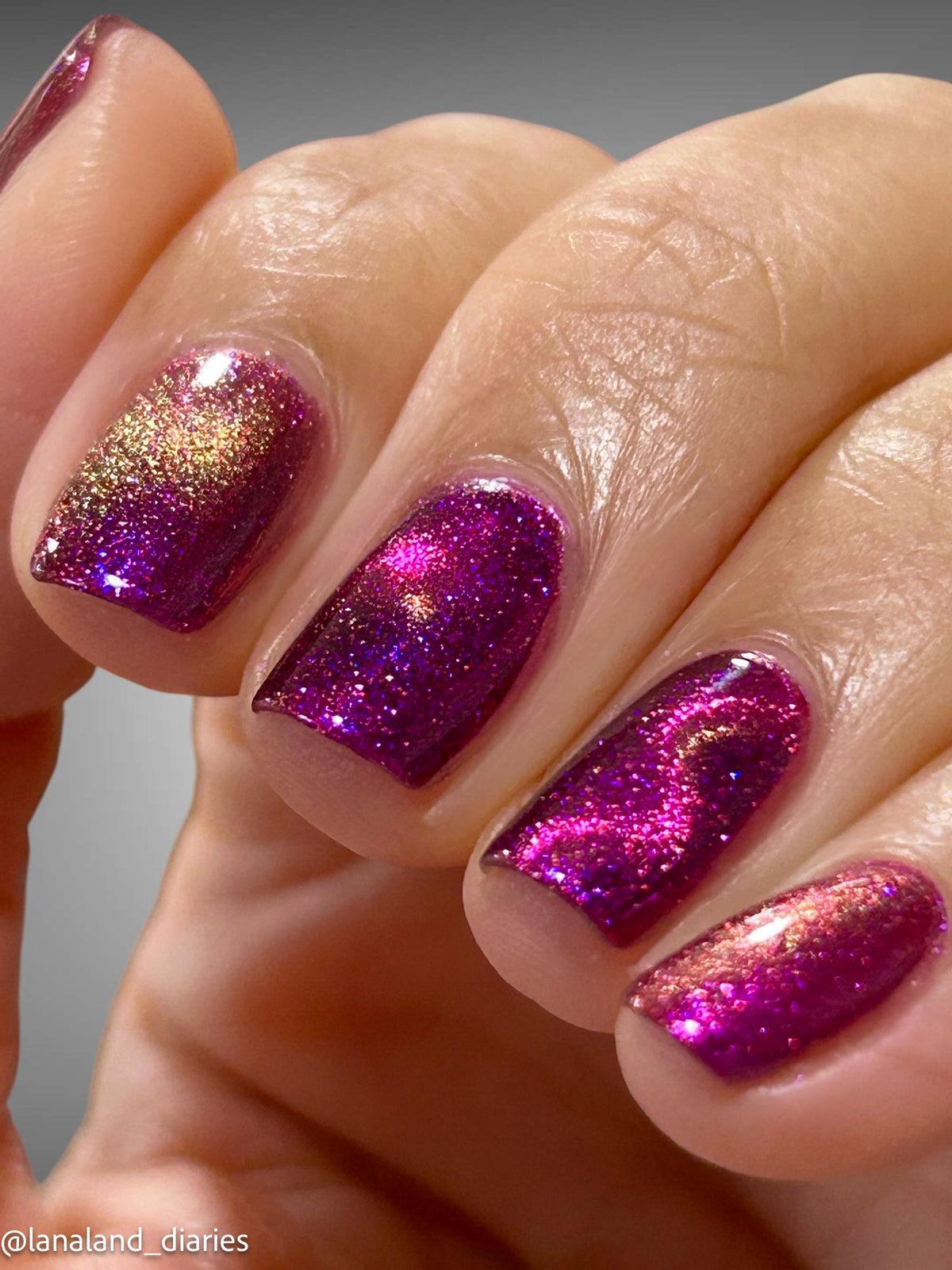 Close-up of a hand with fingernails painted in a sparkling magenta-purple glitter polish with a gold reflective shimmer, against a neutral gray background.