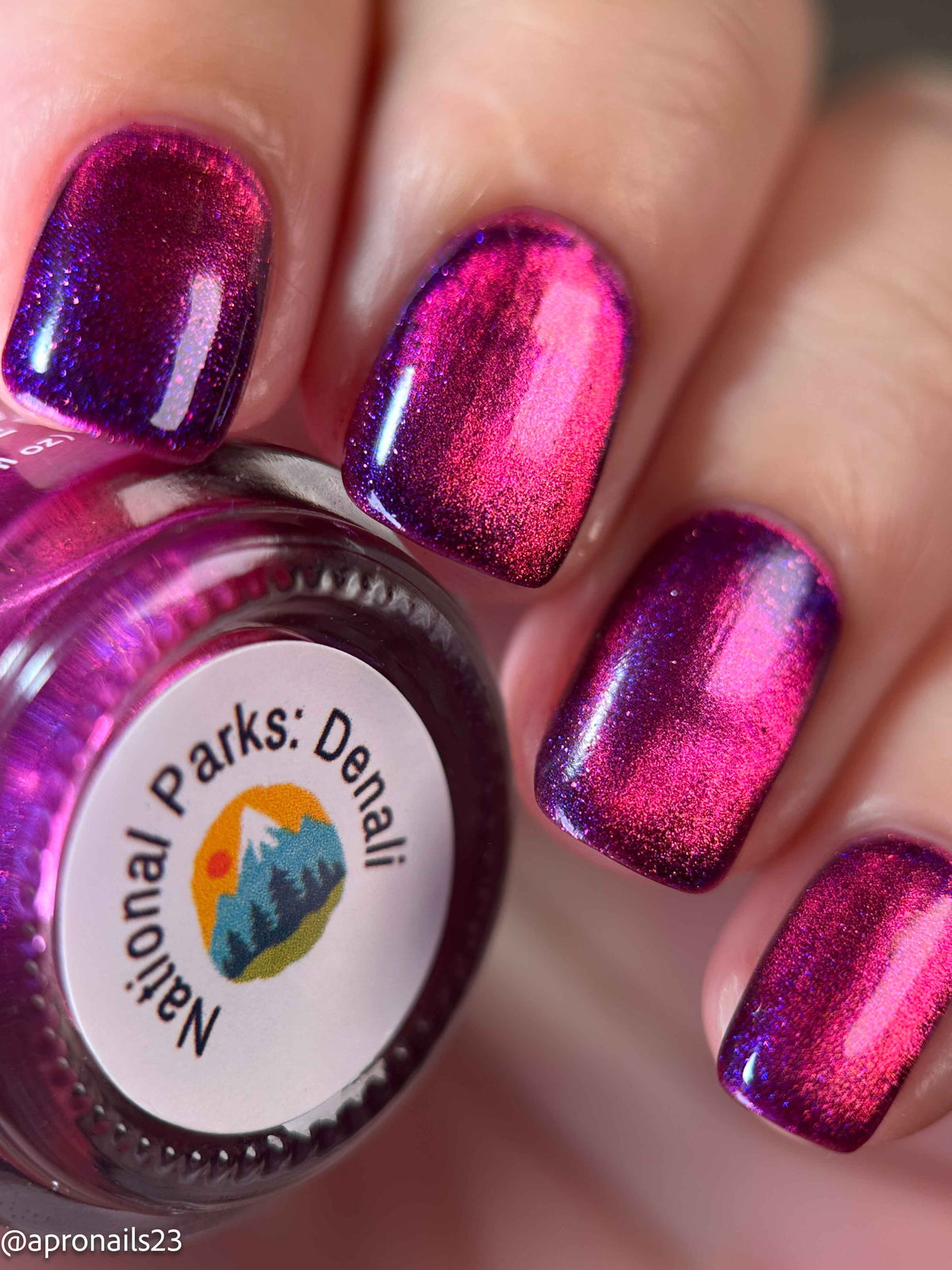 Close-up of fingernails painted with a shimmering metallic magenta-pink and purple polish, with a bottle labeled National Parks: Denali partially visible in the lower left.
