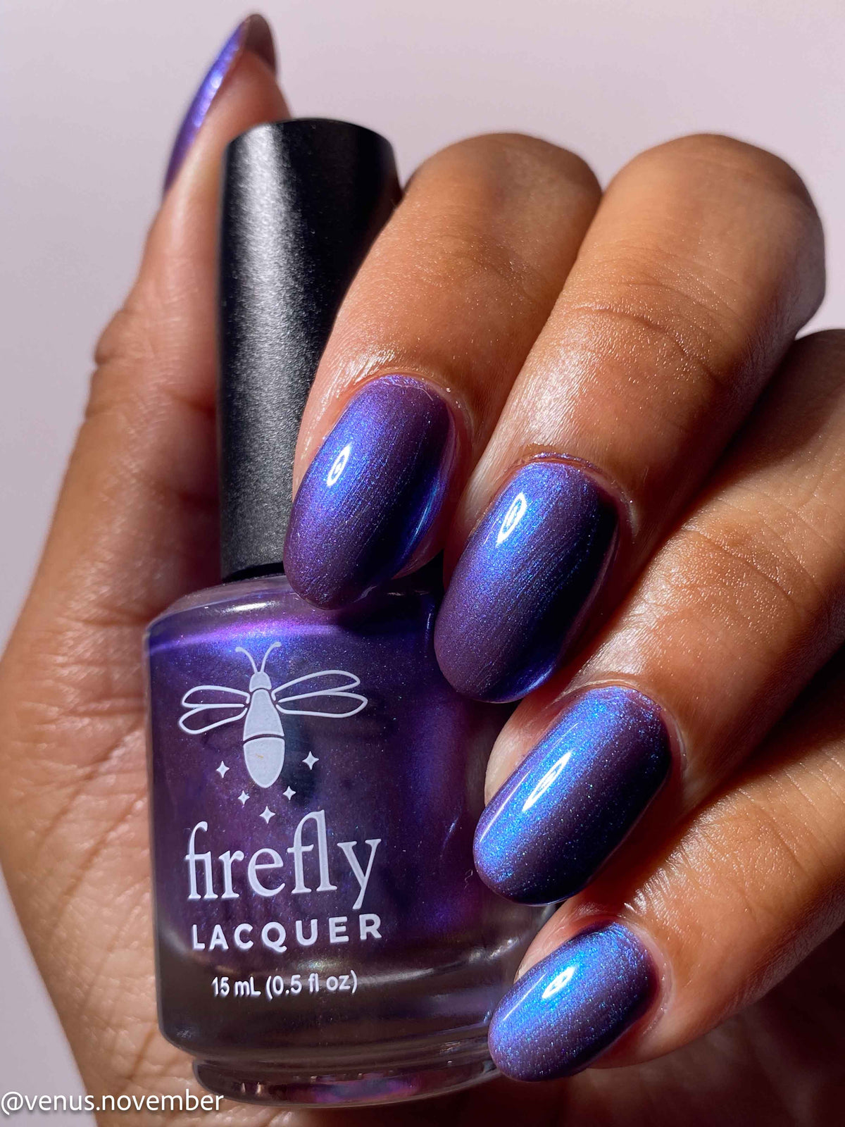 A hand holding a bottle of Firefly Lacquer nail polish, showing the purple and blue iridescent polish applied to the fingernails against a light pink background.
