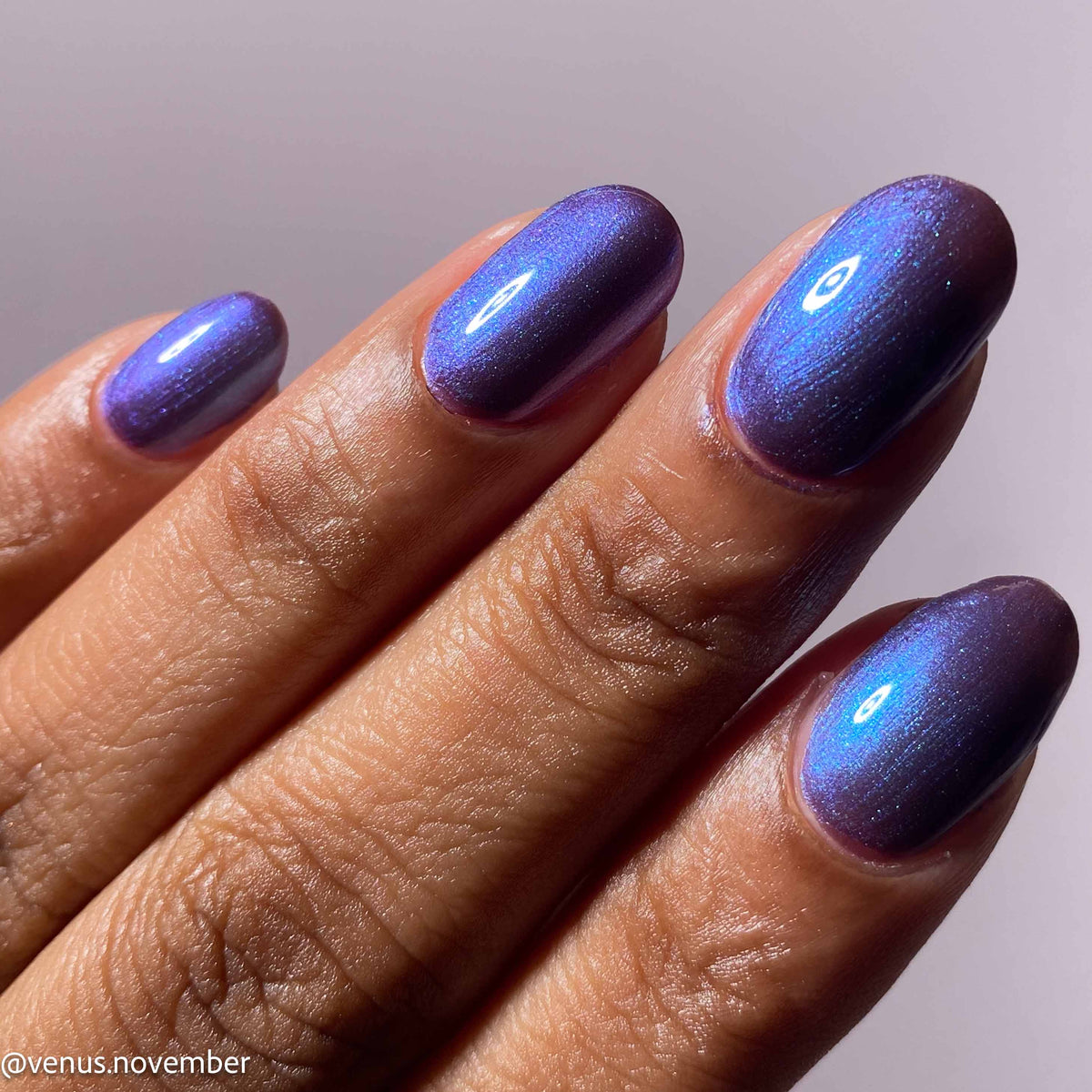 Close-up of a hand with manicured nails painted in a shimmering metallic purple polish that shifts blue under light, shown against a soft neutral background.