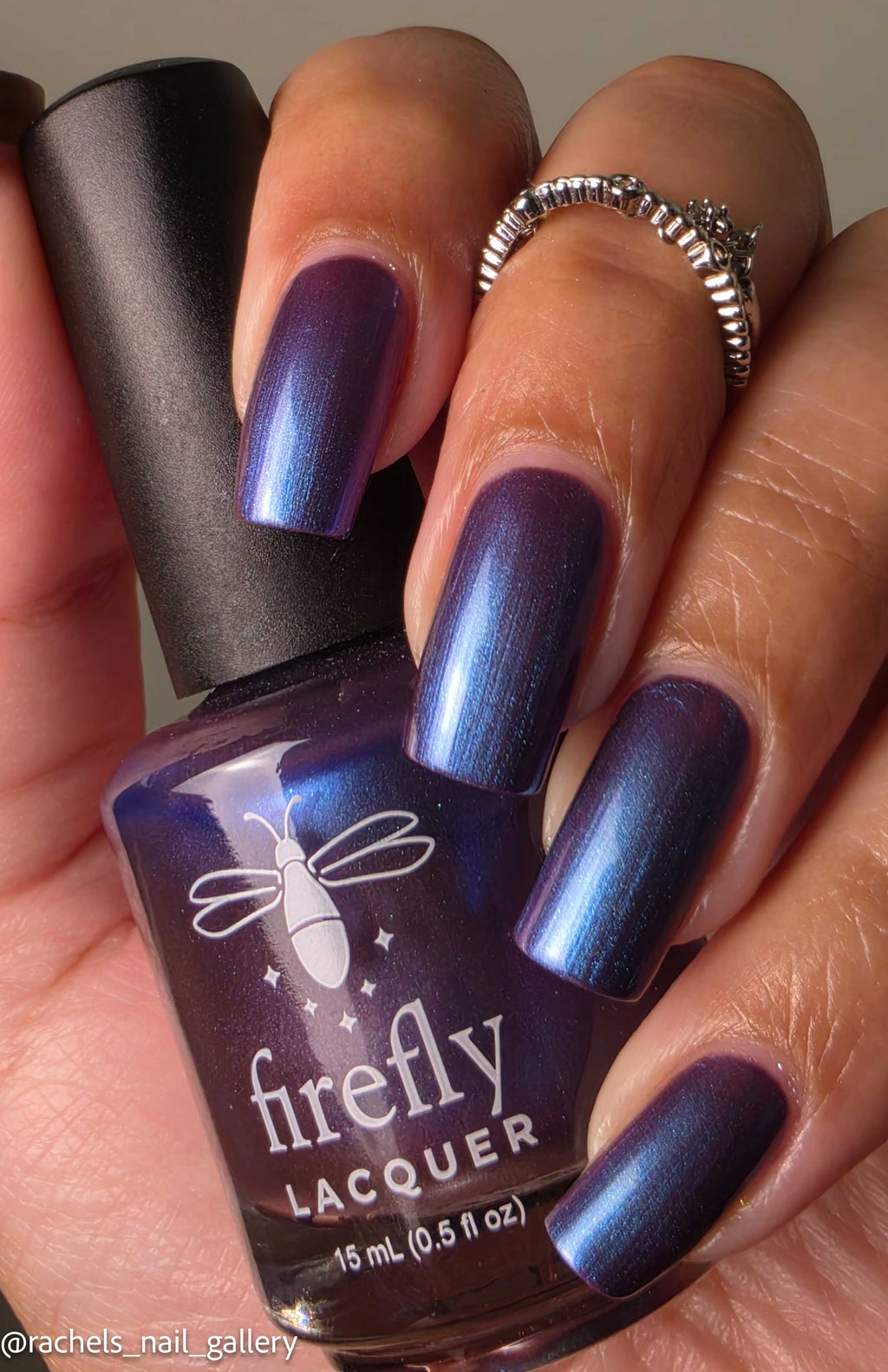 A close-up of a hand with nails painted in a shimmering metallic purple-blue lacquer, holding a bottle of Firefly Lacquer. The bottle label clearly shows the brand name 'firefly LACQUER'.