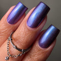 Close-up of a hand with long, almond-shaped nails painted in a shimmery, iridescent purple polish that shifts between blue, violet, and pink. The hand is wearing stacked silver rings against a neutral gray background.