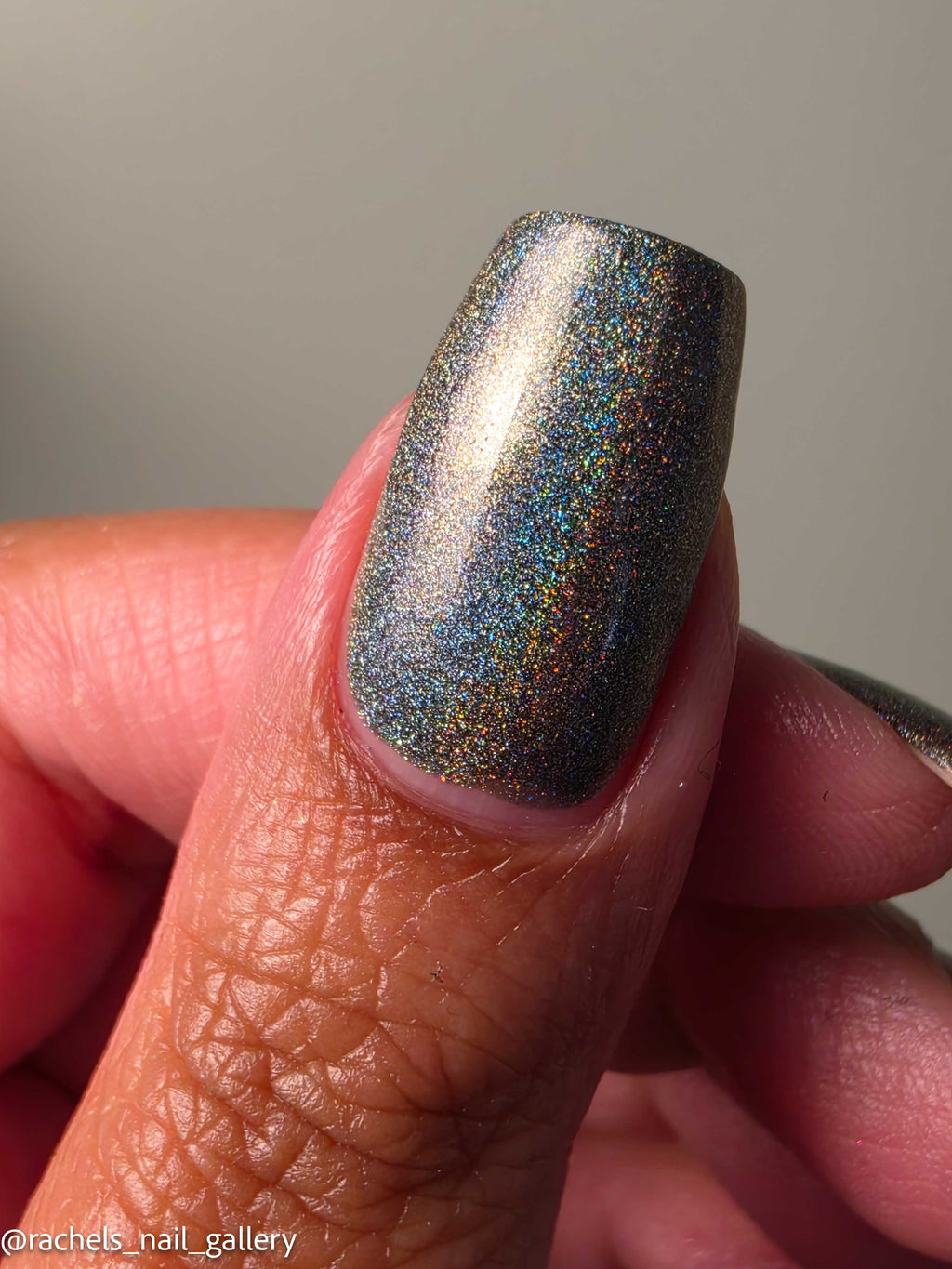 A close-up of a fingernail painted with a holographic glitter nail polish that shifts between gray, blue, and gold depending on the angle, held by a hand against a neutral gray background.
