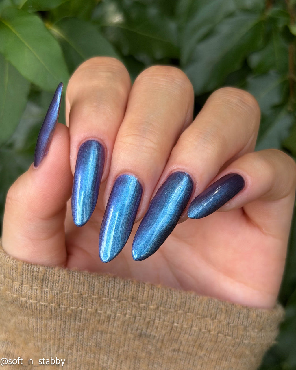A close-up of a hand with long, almond-shaped nails painted in a metallic blue-to-purple iridescent nail polish, set against a background of green leaves.