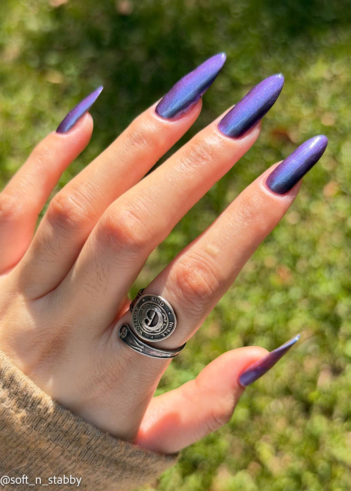 A close-up of a hand with long, metallic purple-to-blue ombre almond-shaped nails, wearing a silver ring, set against a blurred green grass background.