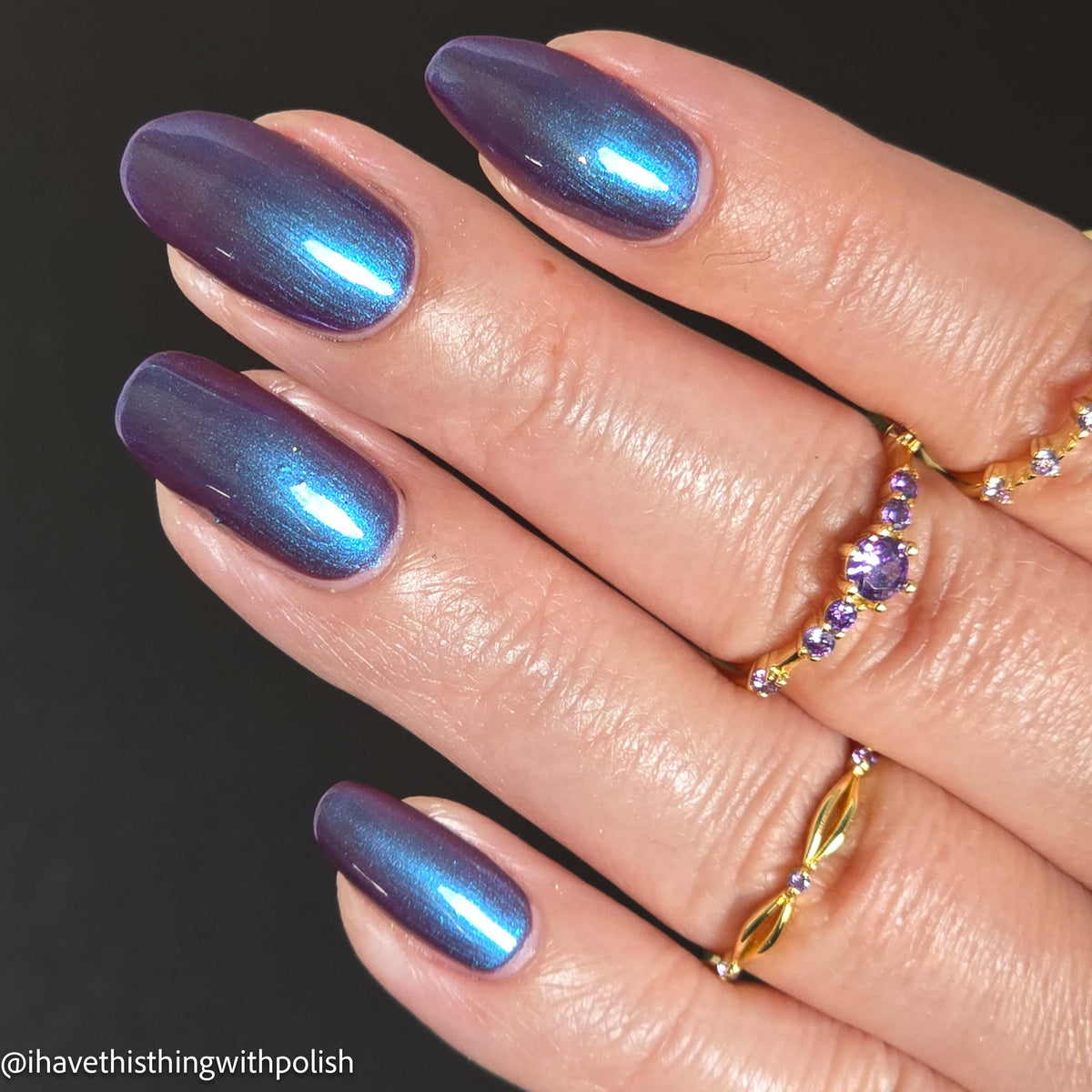 A close-up of a hand with long almond-shaped nails painted in a metallic iridescent blue-to-purple polish, wearing gold rings against a dark background.