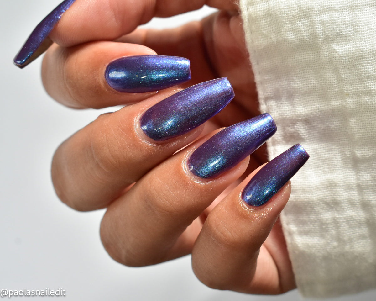 A close-up of a hand with long coffin-shaped nails painted in a shimmery iridescent purple-to-blue nail polish, with a white textured sleeve visible on the right side.