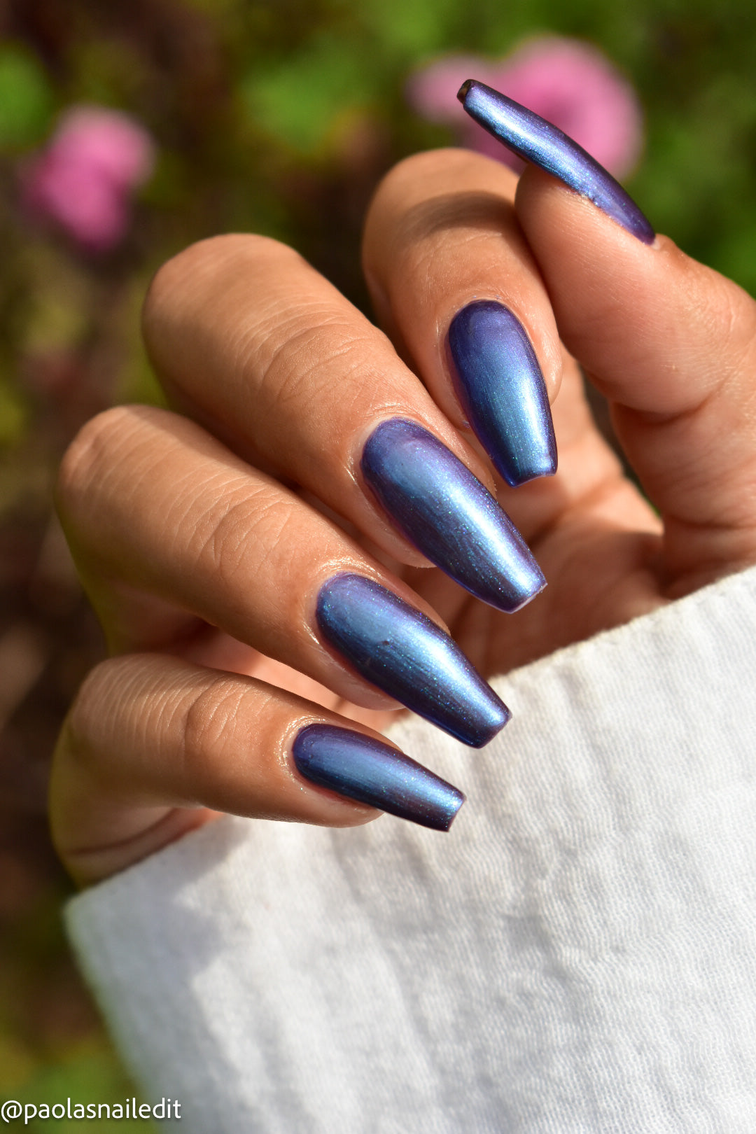 A close-up of a hand with long, almond-shaped nails painted in a metallic, iridescent blue polish that shifts from deep navy to purple. The background is a soft, blurred outdoor garden with green foliage and pink flowers.
