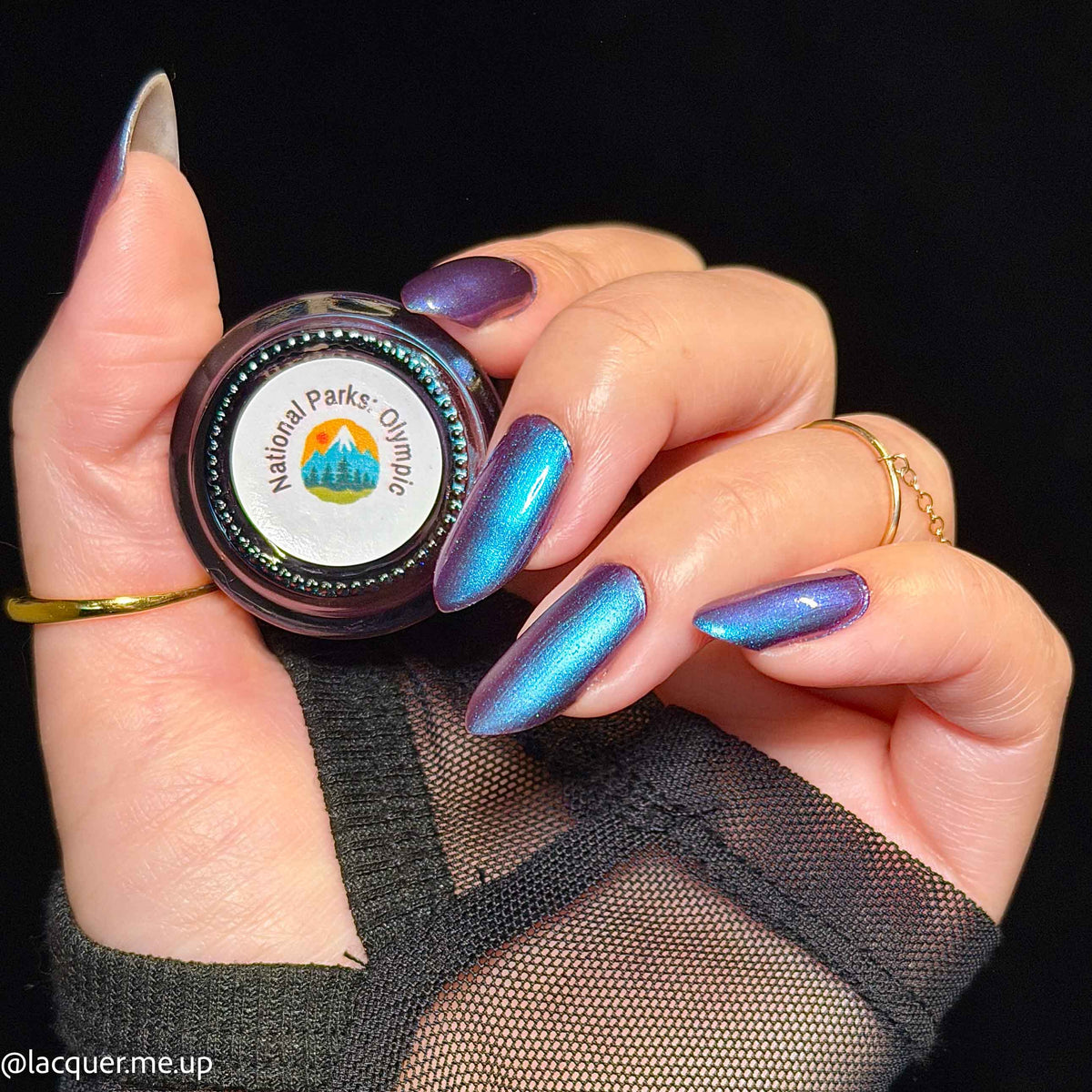 A close-up of a hand with long, metallic iridescent nails painted in a blue-to-purple shift, holding a small round jar labeled National Parks Olympus. The background is solid black.
