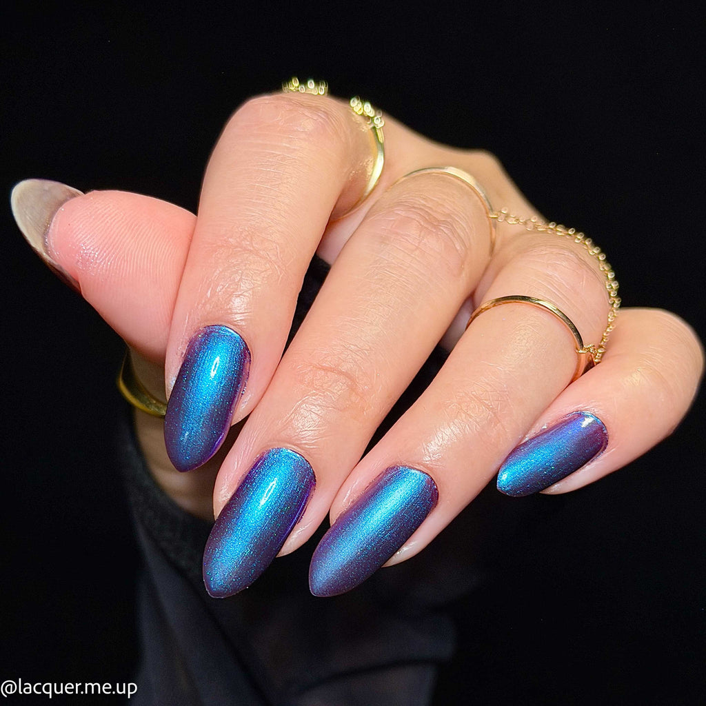 A close-up of a hand with almond-shaped nails painted in a shimmering metallic blue-to-purple ombre nail polish, styled with gold rings against a black background.