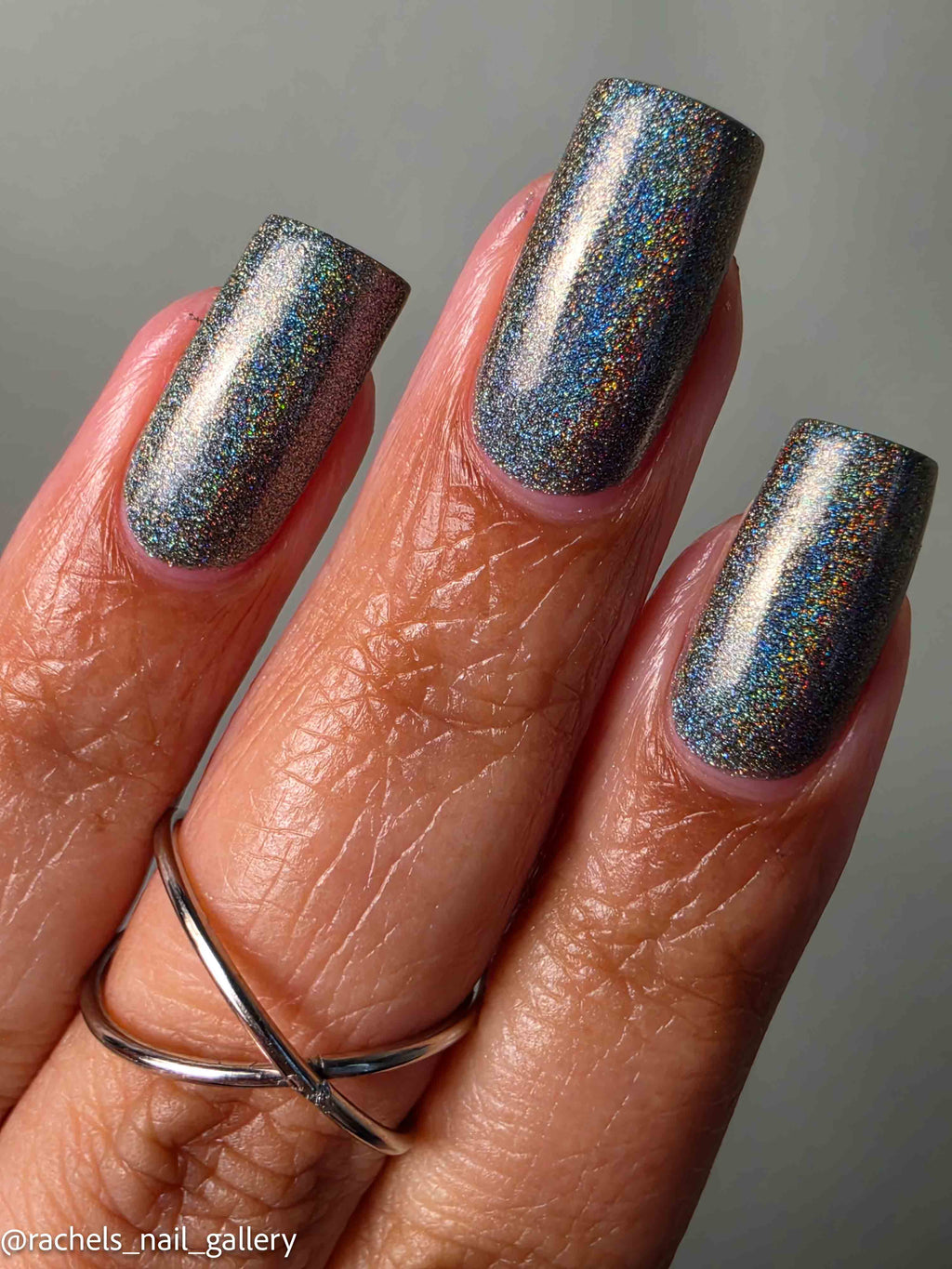 A close-up of a hand with long, almond-shaped nails painted in a shimmering holographic nail polish that reflects silver, blue, and gold tones. A silver ring is worn on the ring finger against a neutral gray background.