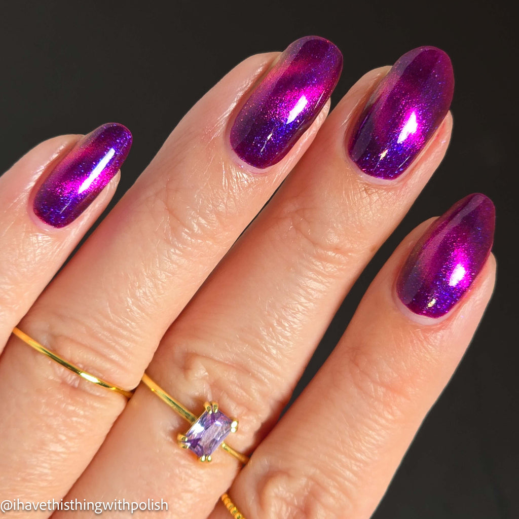 A close-up of a hand with long almond-shaped nails painted in a metallic magenta-to-purple iridescent nail polish, wearing two thin gold rings against a dark background.