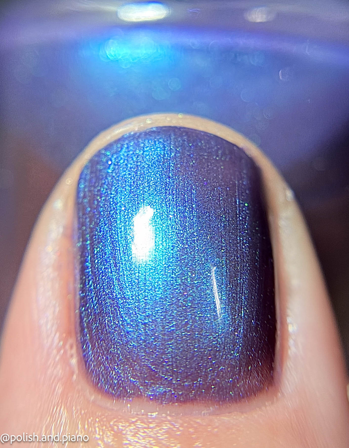A close-up of a fingernail painted with a shimmering iridescent blue-purple nail polish that shifts with light, set against a blurred matching blue background.