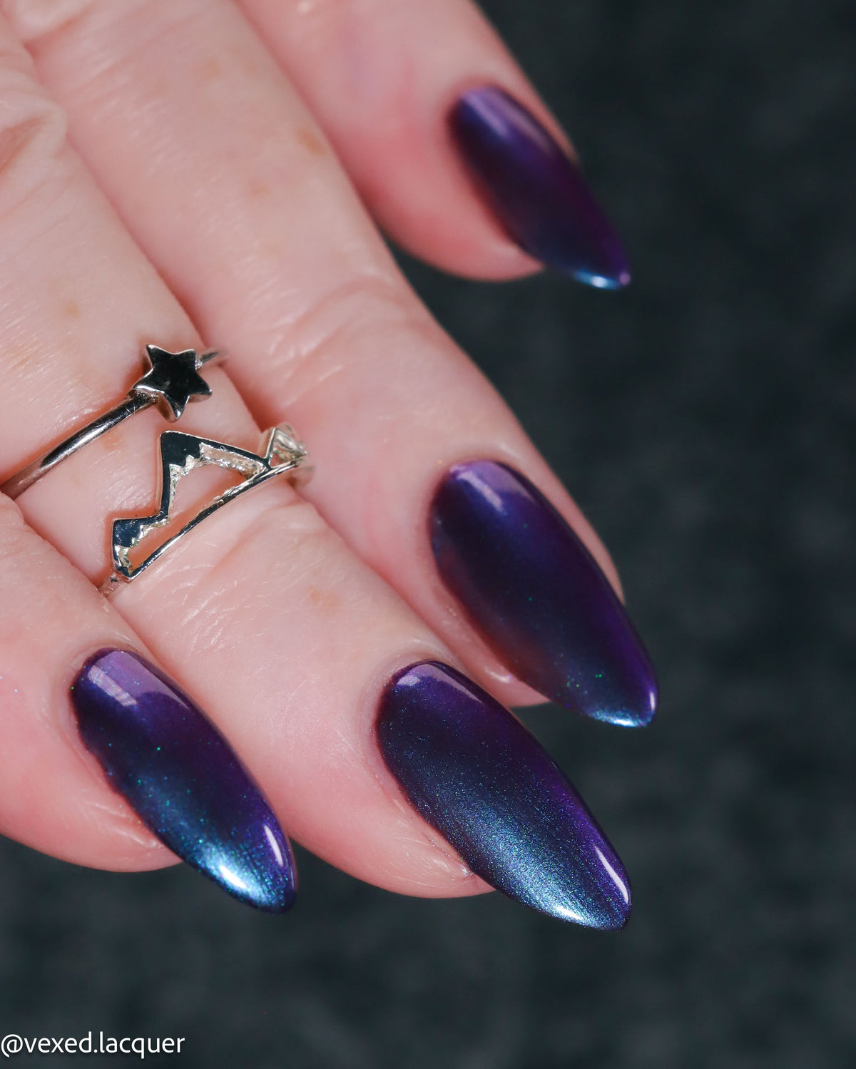 A close-up of a hand with long almond-shaped nails painted in a metallic ombre gradient shifting from deep purple to blue, wearing a silver ring with a black star on the ring finger. The background is a dark, textured gray surface.