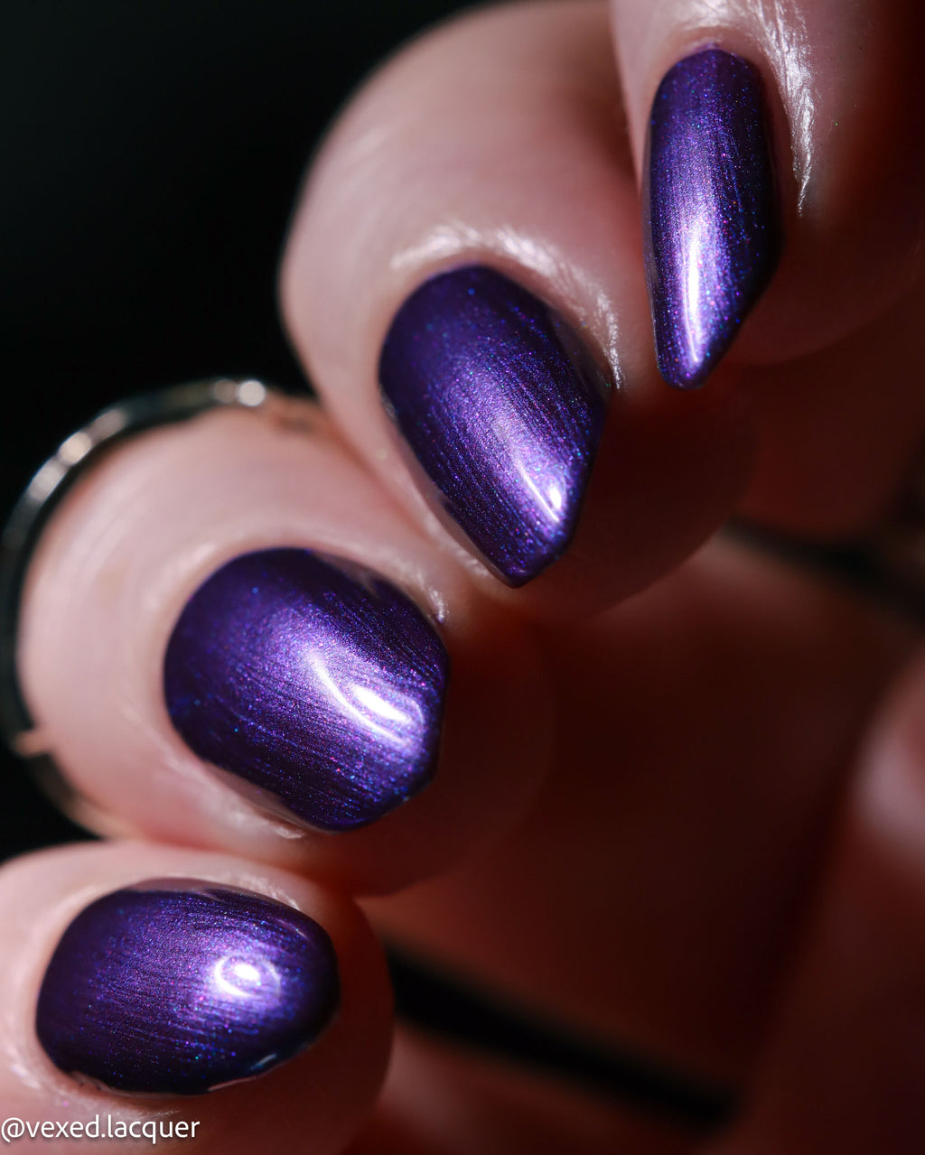 Close-up of a hand with fingernails painted in a metallic, shimmering deep purple lacquer. A silver ring is visible on the left finger against a dark, out-of-focus background.
