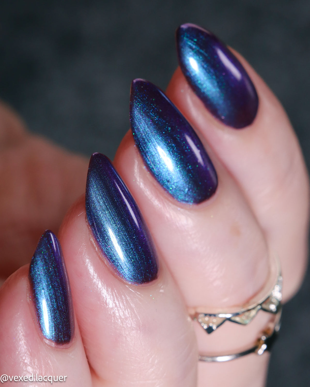 Close-up of a hand with long almond-shaped nails painted in a shimmering metallic blue-to-purple nail polish, with silver rings on one finger against a dark gray background.