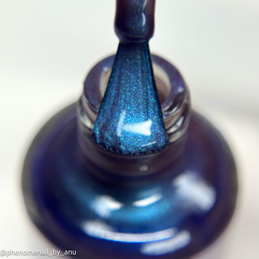 Close-up of a blue metallic nail polish brush stroke applied to a glass bottle, showing a shimmering deep blue color with fine glitter.