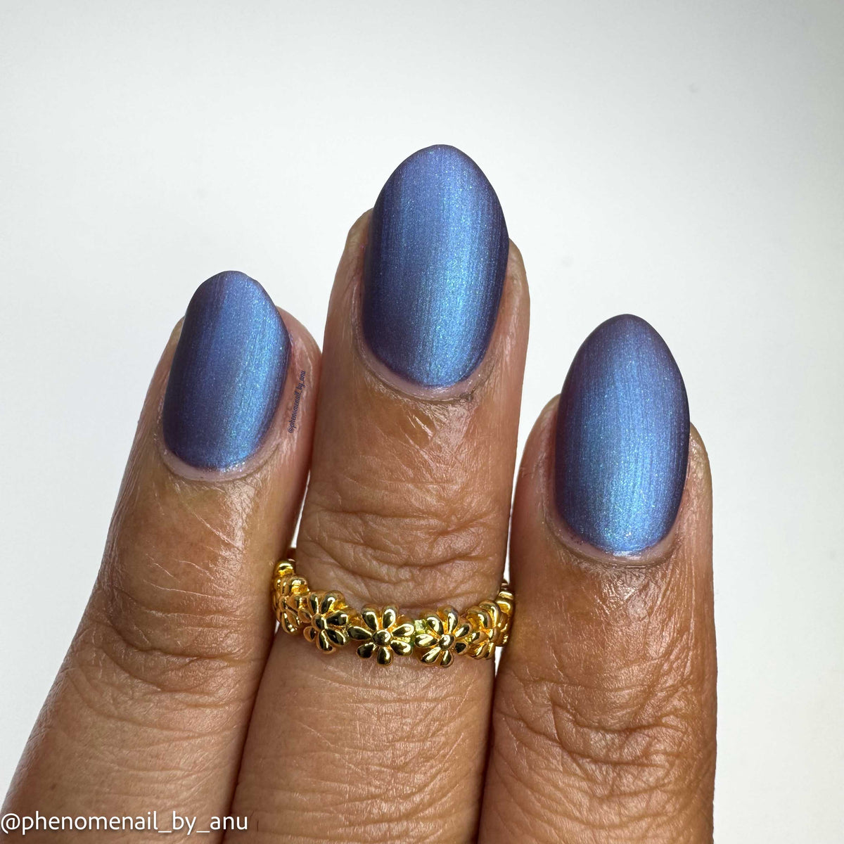 A close-up of a hand with manicured nails painted in a shimmery periwinkle blue shade, wearing a gold flower ring. The watermark in the bottom-left corner reads '@phenomenail_by_anu'.