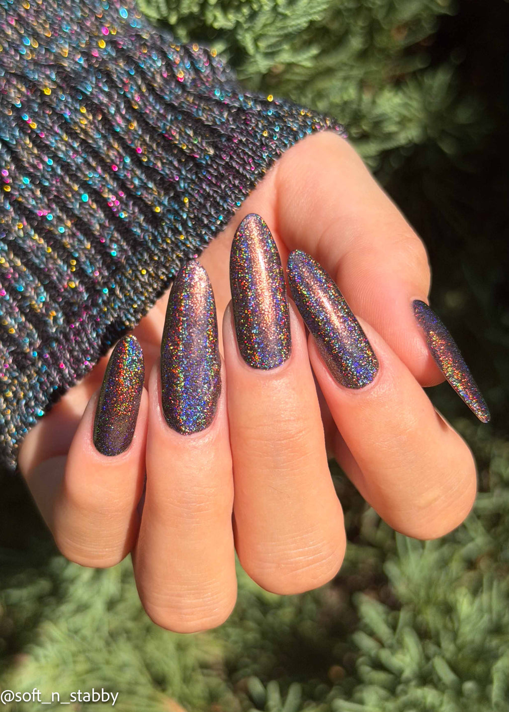 A close-up of a hand with long, almond-shaped nails painted in a shimmering holographic glitter polish that shifts between purple, blue, and pink. The hand is wearing a dark ribbed knit sweater sleeve, with a blurred green outdoor background.