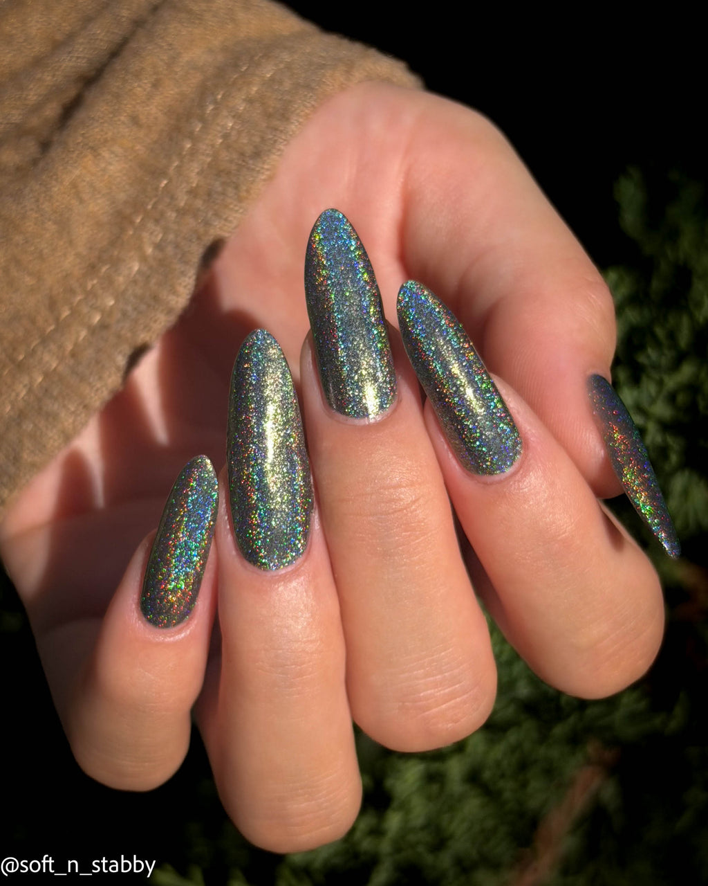 A close-up of a hand with long almond-shaped nails painted in a holographic glitter nail polish that shifts between blue, green, and gold, shown against a dark outdoor background.