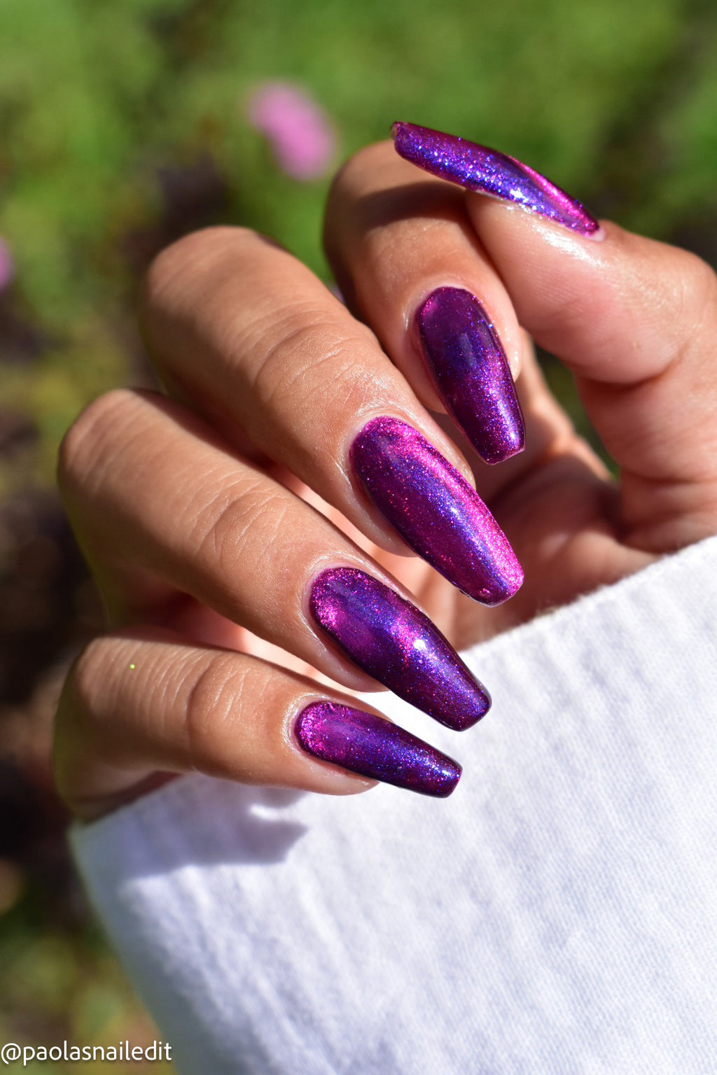 A close-up of a hand with long, almond-shaped nails painted in a shimmering metallic purple and pink nail polish, set against a blurred green outdoor background.
