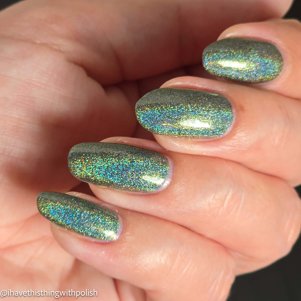 Close-up of a hand with fingernails painted in a shimmering holographic nail polish that shifts between green, blue, and gold tones.