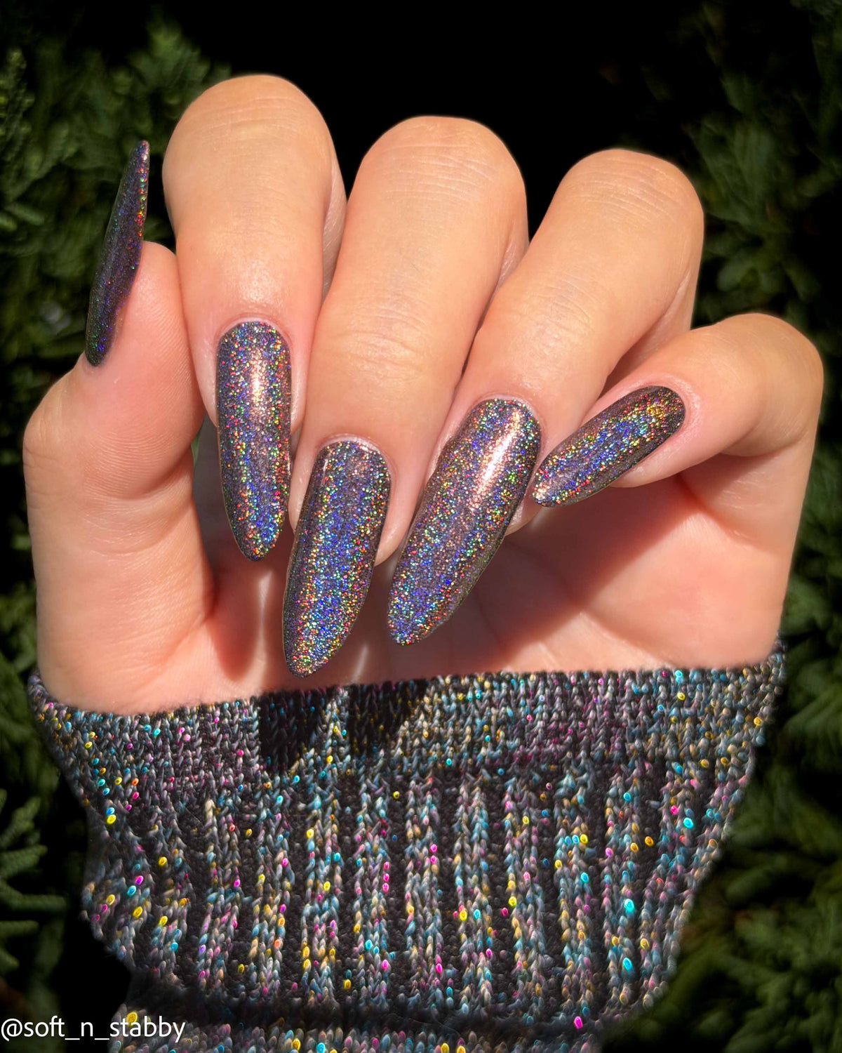 A close-up of a hand with long almond-shaped nails painted in a shimmering holographic glitter nail polish, paired with a ribbed knit sweater featuring multicolor flecks, set against a dark green pine background.