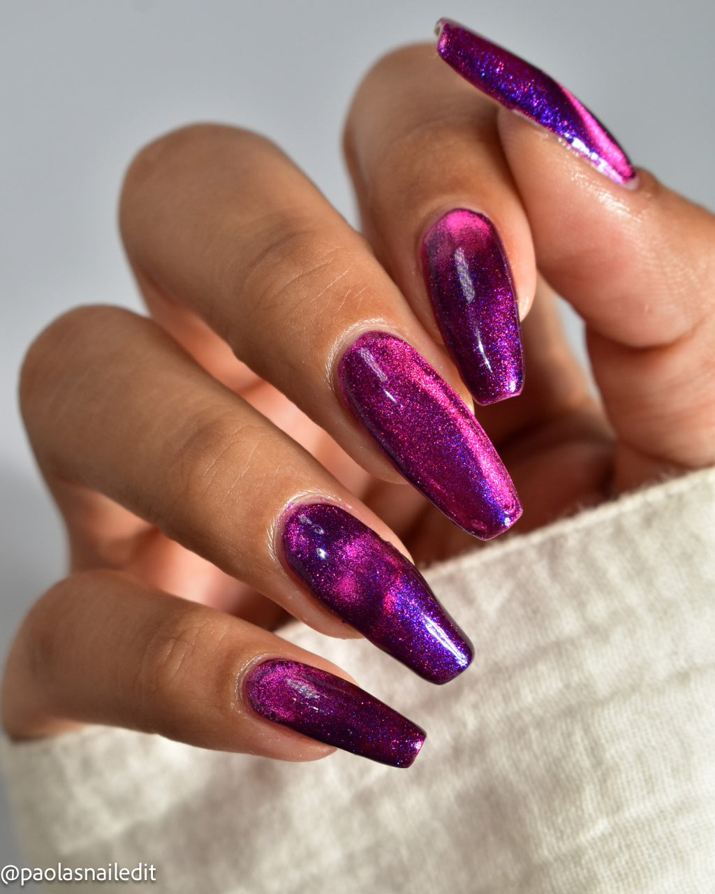 A close-up of a hand with long coffin-shaped nails painted in a sparkling metallic magenta and purple gradient nail polish, resting against a light beige textured fabric.