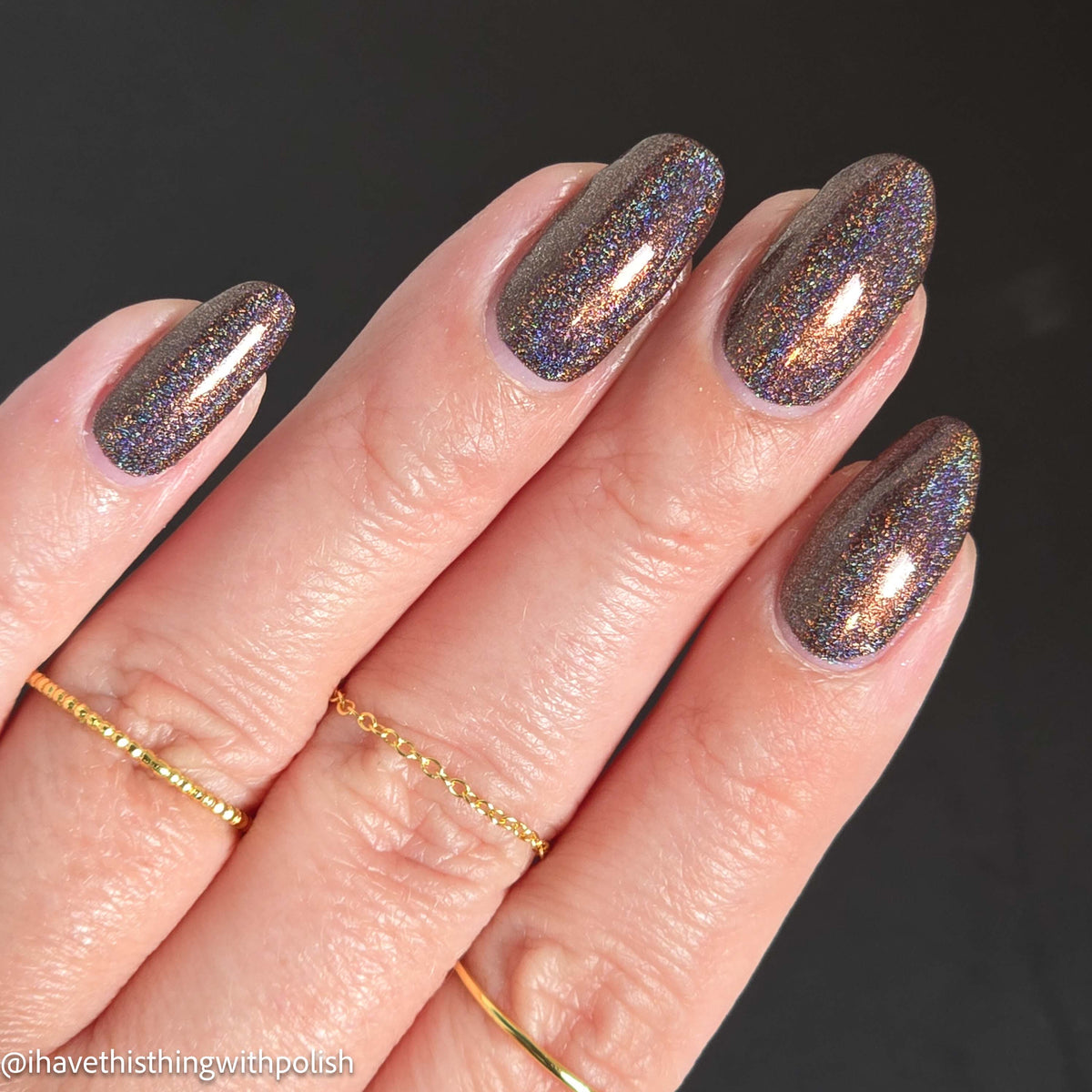 A close-up of a hand with almond-shaped nails painted in a dark shimmery nail polish with holographic iridescent glitter. The nails reflect purple, blue, and gold tones against a dark background.