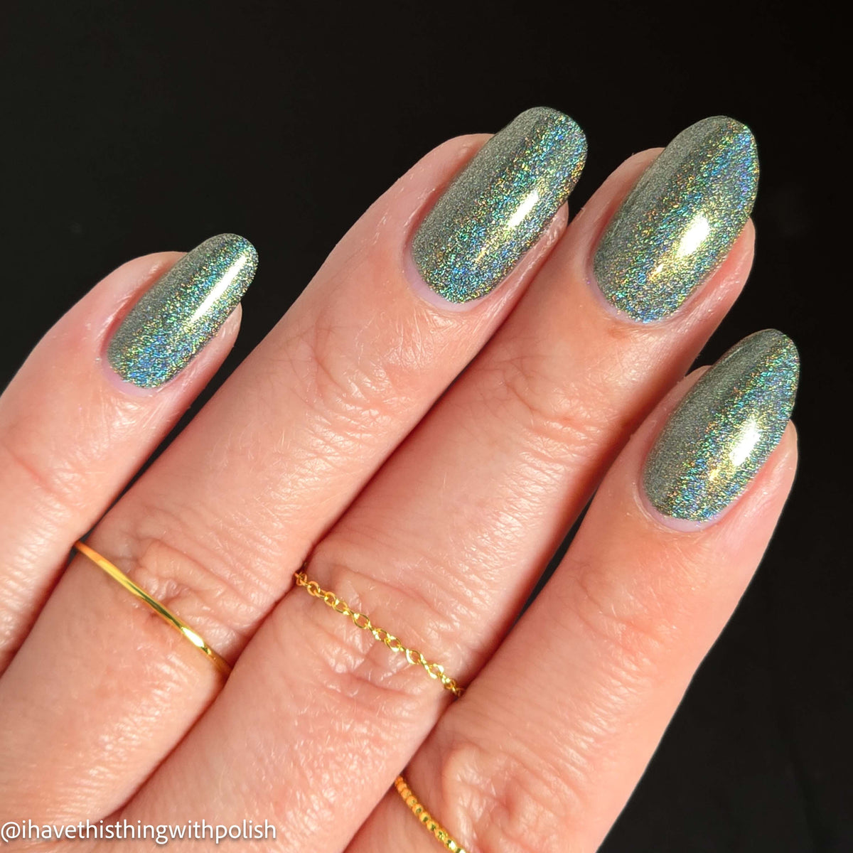A close-up of a hand with almond-shaped nails painted in a shimmering holographic nail polish that shifts between blue, green, and gold. The hand is adorned with three delicate gold rings against a solid black background.