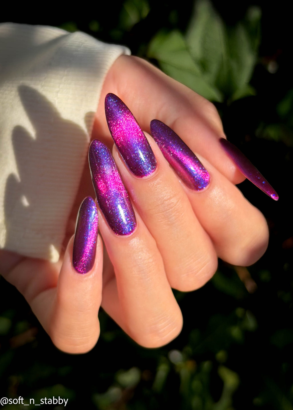 A close-up of a hand with long, stiletto-shaped nails painted in a shimmering holographic purple and pink polish, set against a dark green outdoor background.
