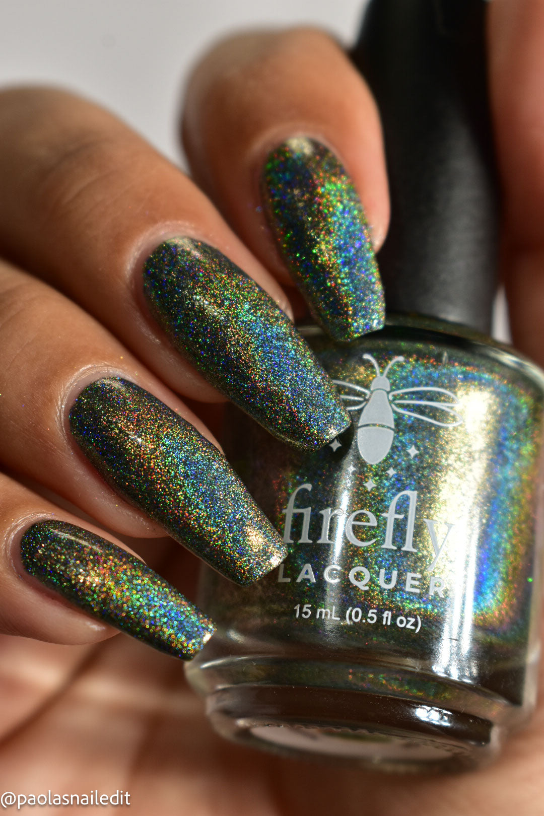 A close-up of a hand with long almond-shaped nails painted in a shimmering holographic nail polish, holding a bottle of Firefly Lacquer in the same shade. The polish reflects green, blue, and gold iridescent glitter.