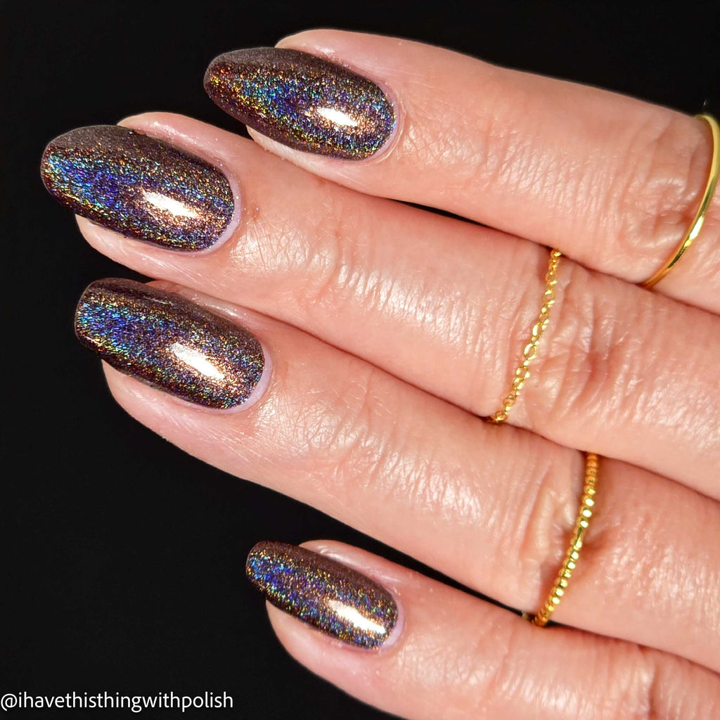 Close-up of a hand with manicured nails painted in a dark brown base with dense iridescent holographic glitter that shifts between blue, purple, and gold, set against a solid black background.