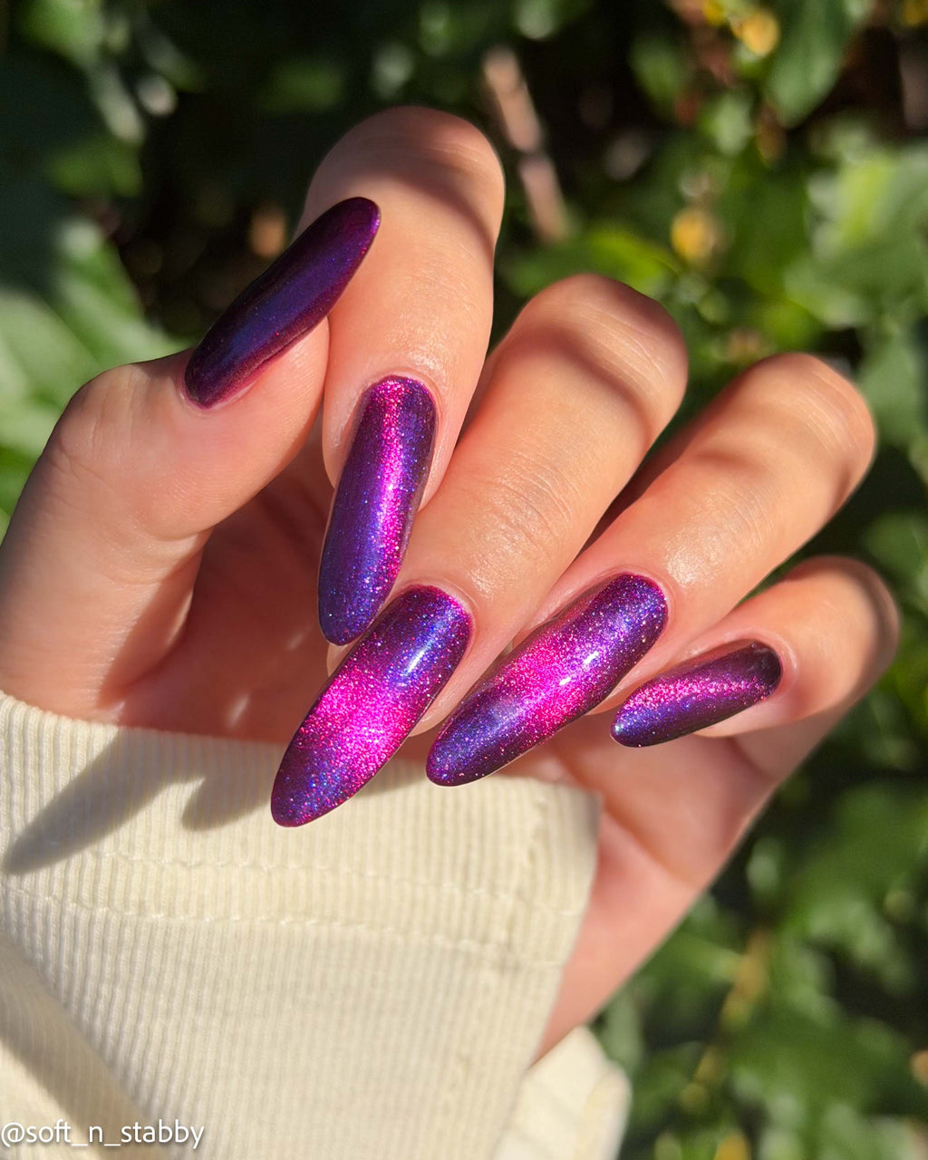 A close-up of a hand with long, almond-shaped nails painted in a shimmering metallic purple-to-pink gradient polish, set against a blurred green outdoor background.