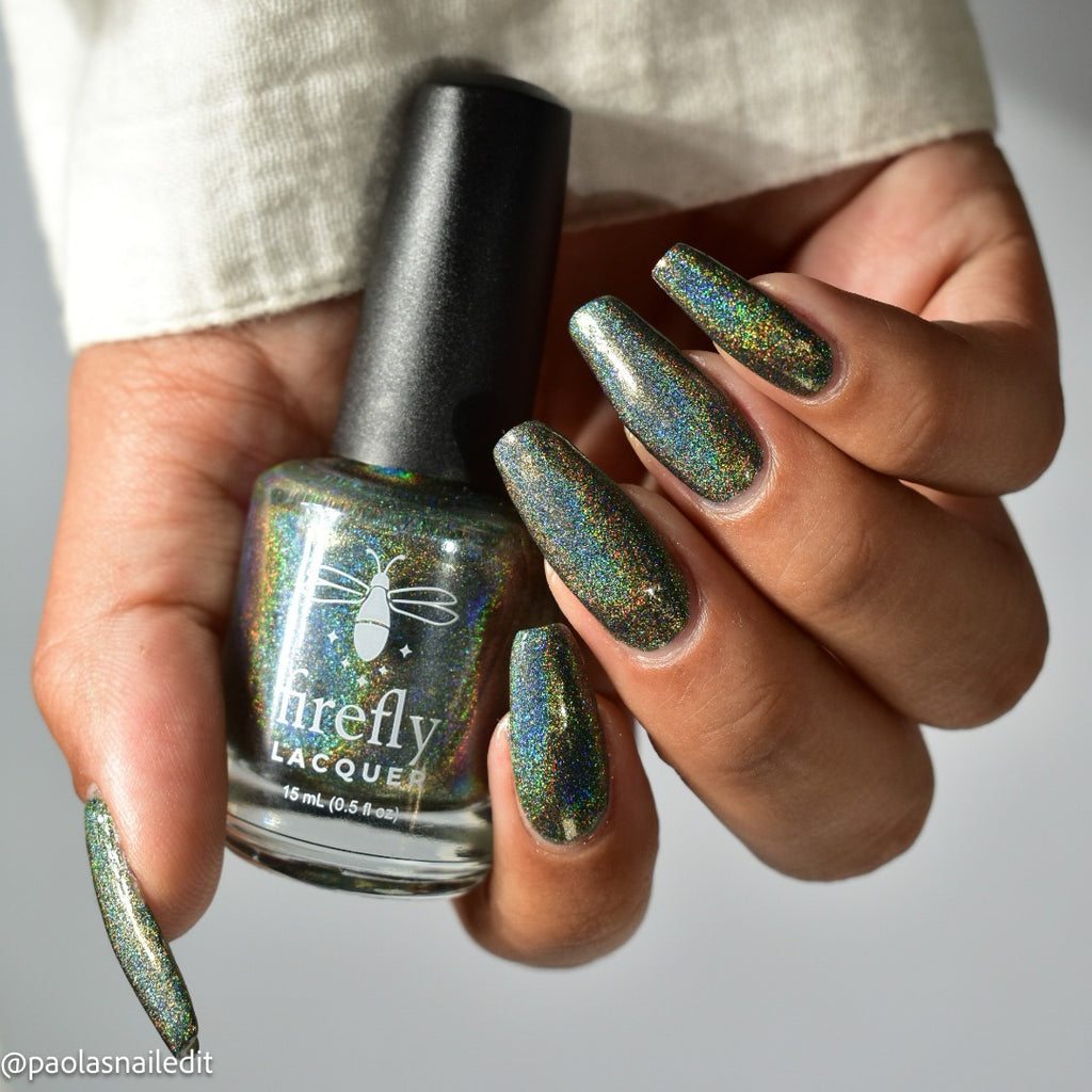A hand holding a bottle of Firefly Lacquer nail polish with matching long, iridescent green-blue manicured nails, against a light gray background.