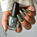 A hand holding a bottle of Firefly Lacquer nail polish with matching long, iridescent green-blue manicured nails, against a light gray background.