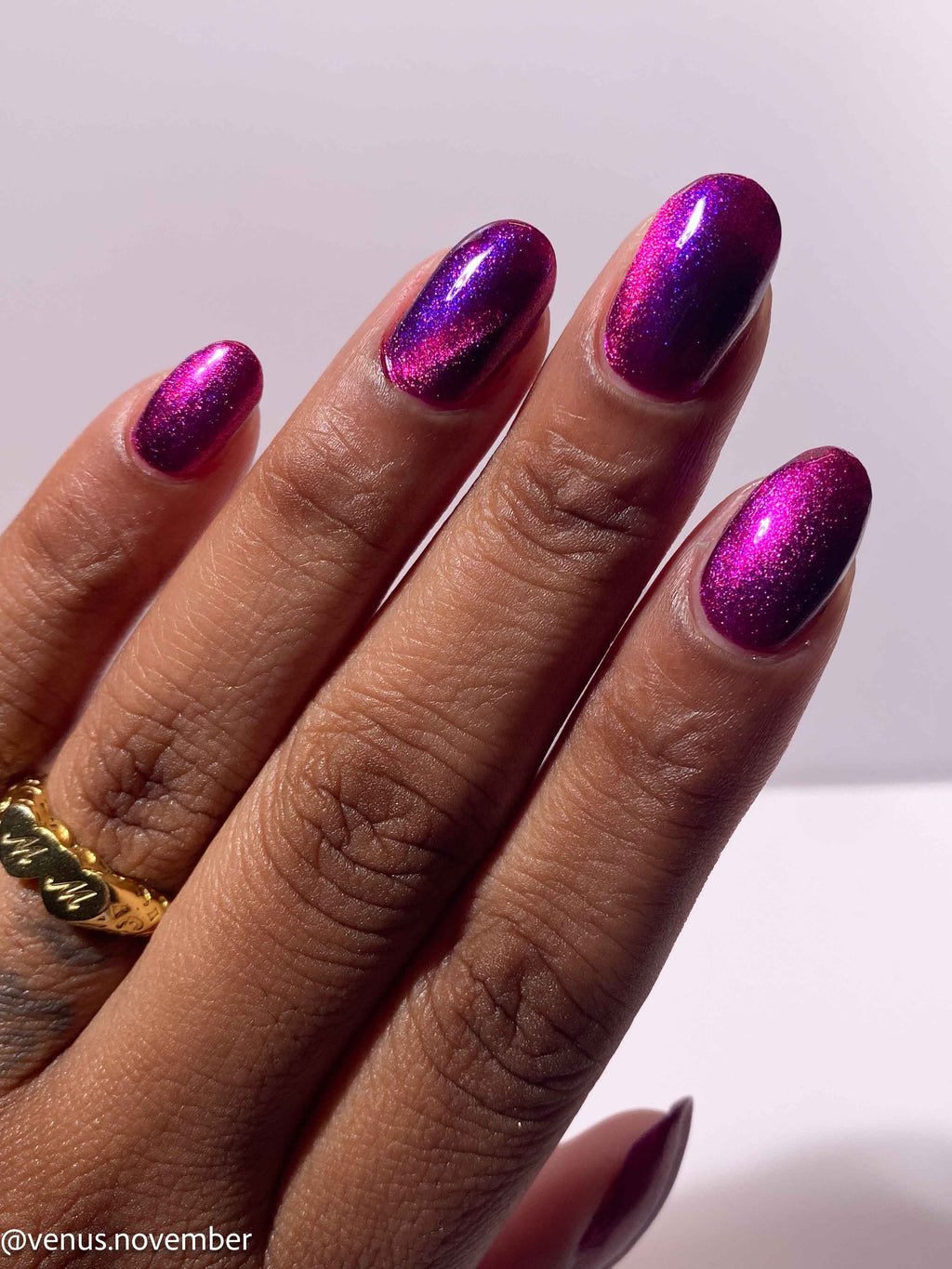 A close-up of a hand with manicured nails painted in a metallic magenta-to-purple gradient polish, wearing a gold ring, against a plain light background.