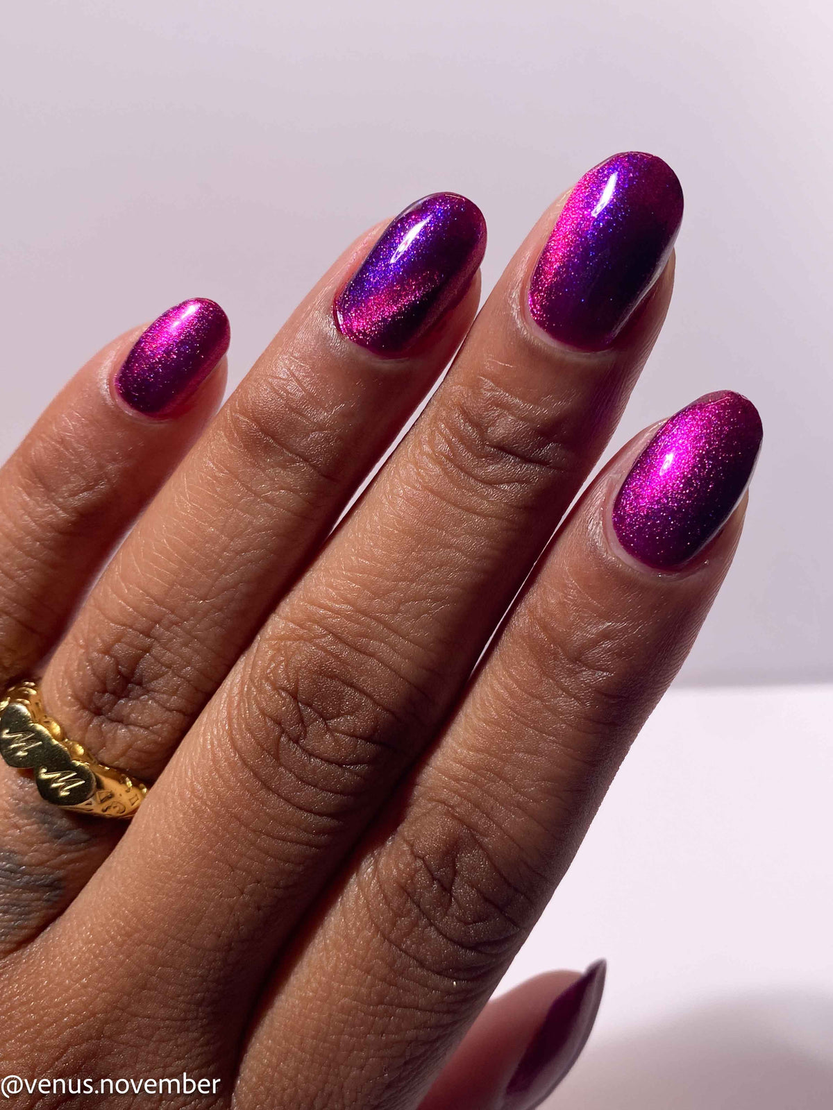 A close-up of a hand with manicured nails painted in a metallic magenta-to-purple gradient polish, wearing a gold ring, against a plain light background.
