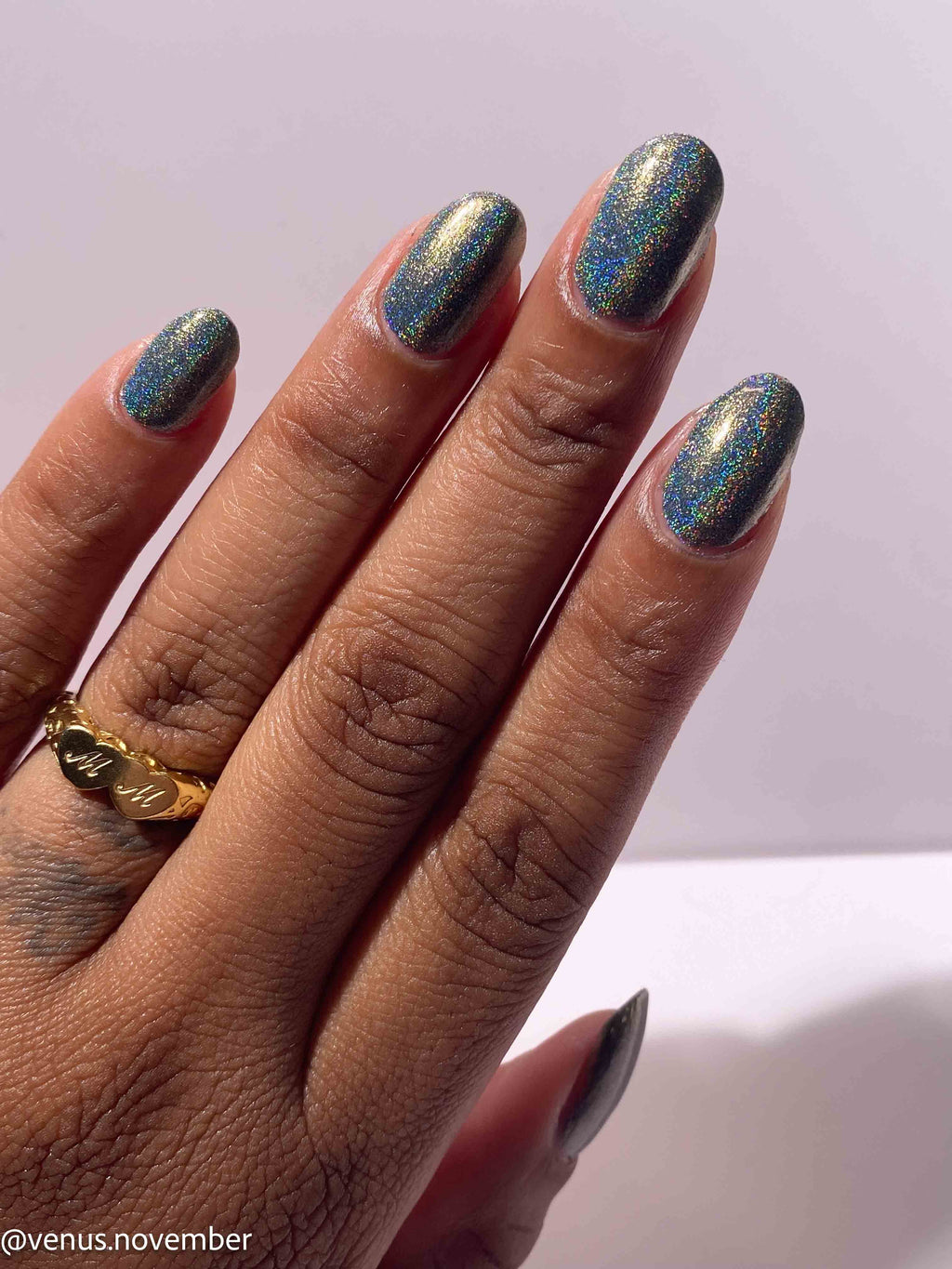 A close-up of a hand with almond-shaped nails painted in a shimmering holographic blue nail polish, showing a gold ring on one finger against a light background.